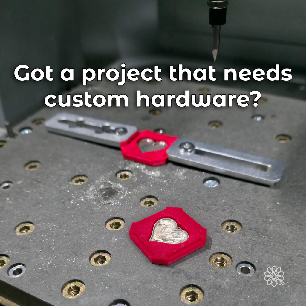 Red 3D-printed CNC fixtures holding two heart-shaped metal pieces on a threaded machine bed beneath an engraving bit, with visible text describing custom hardware projects.