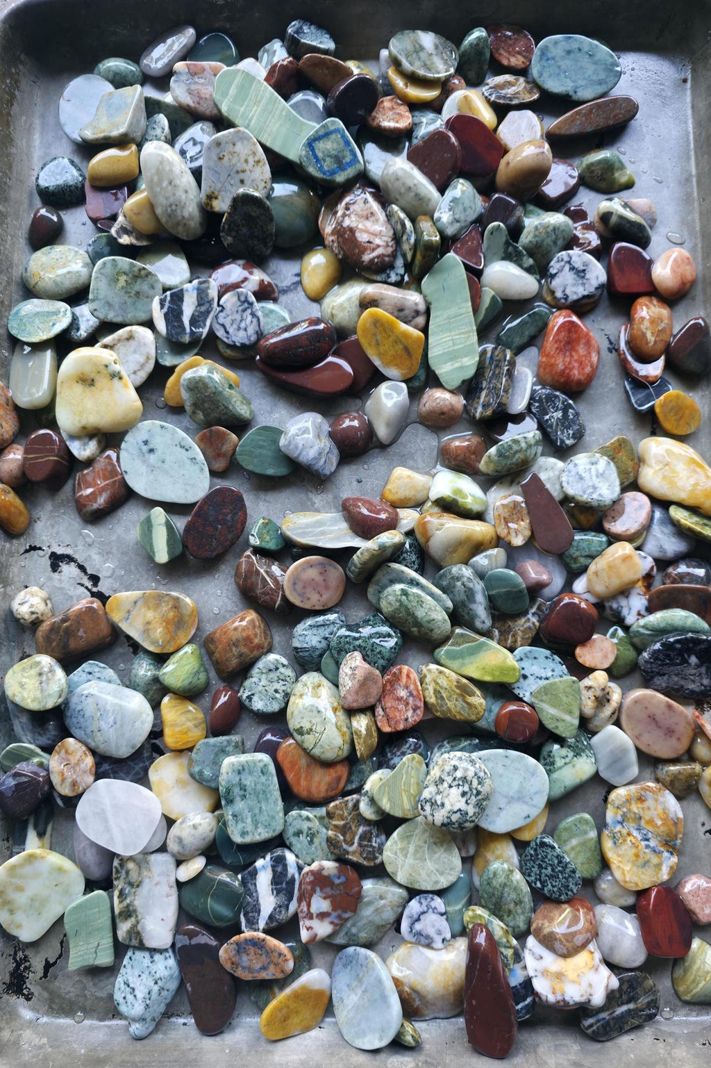 May include: A collection of smooth, polished stones in various colors, and patterns including green, yellow, red, brown, blue and white. The stones are arranged in a single layer.