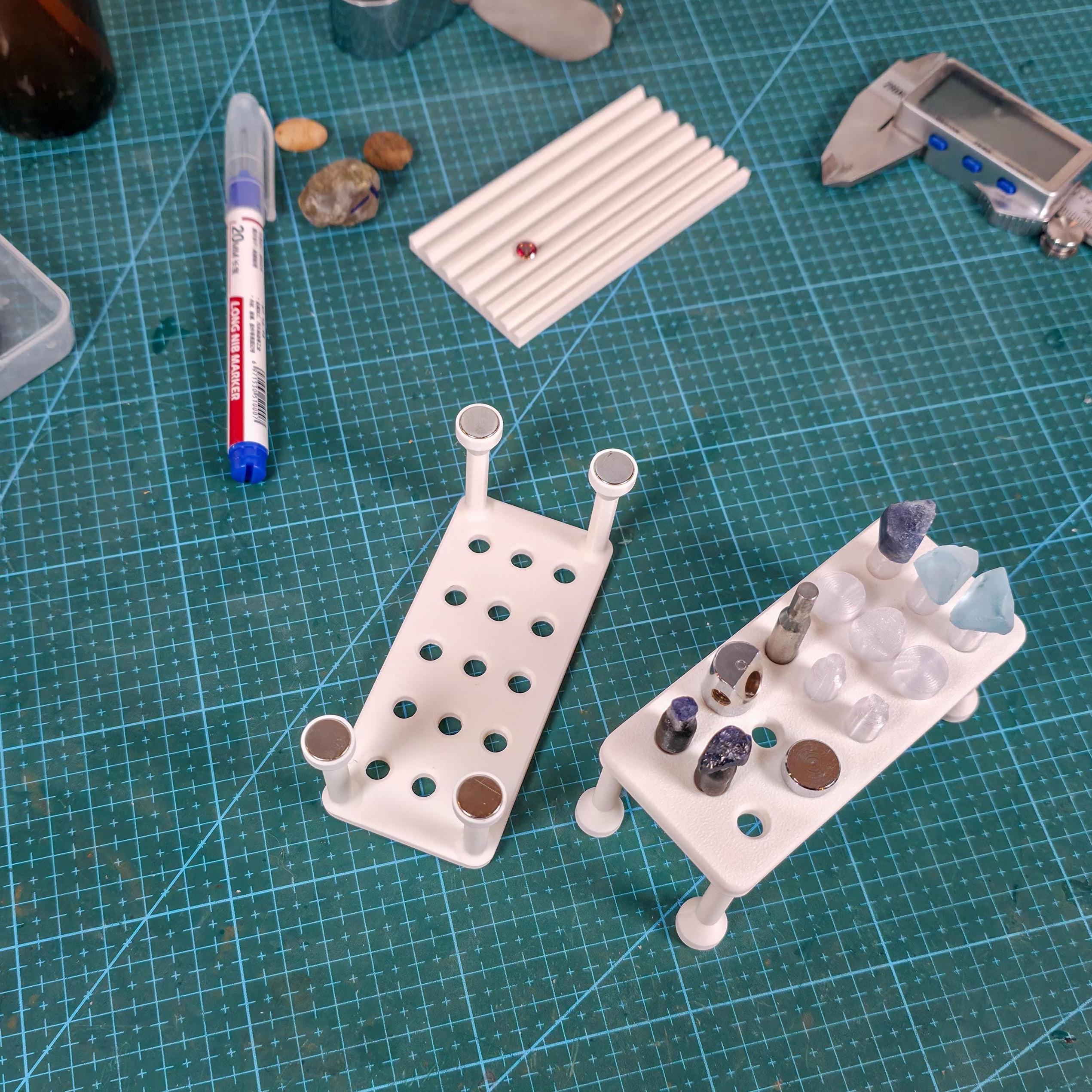 Two magnetic dop trays on a cutting mat holding mixed gemstones and metal dop sticks. The trays keep stones separated, visible, and stable during lapidary and faceting work. Shown in a workshop environment with measuring tools and stones nearby, emphasizing clean workflow organization for cabbing, faceting, and stone preparation.