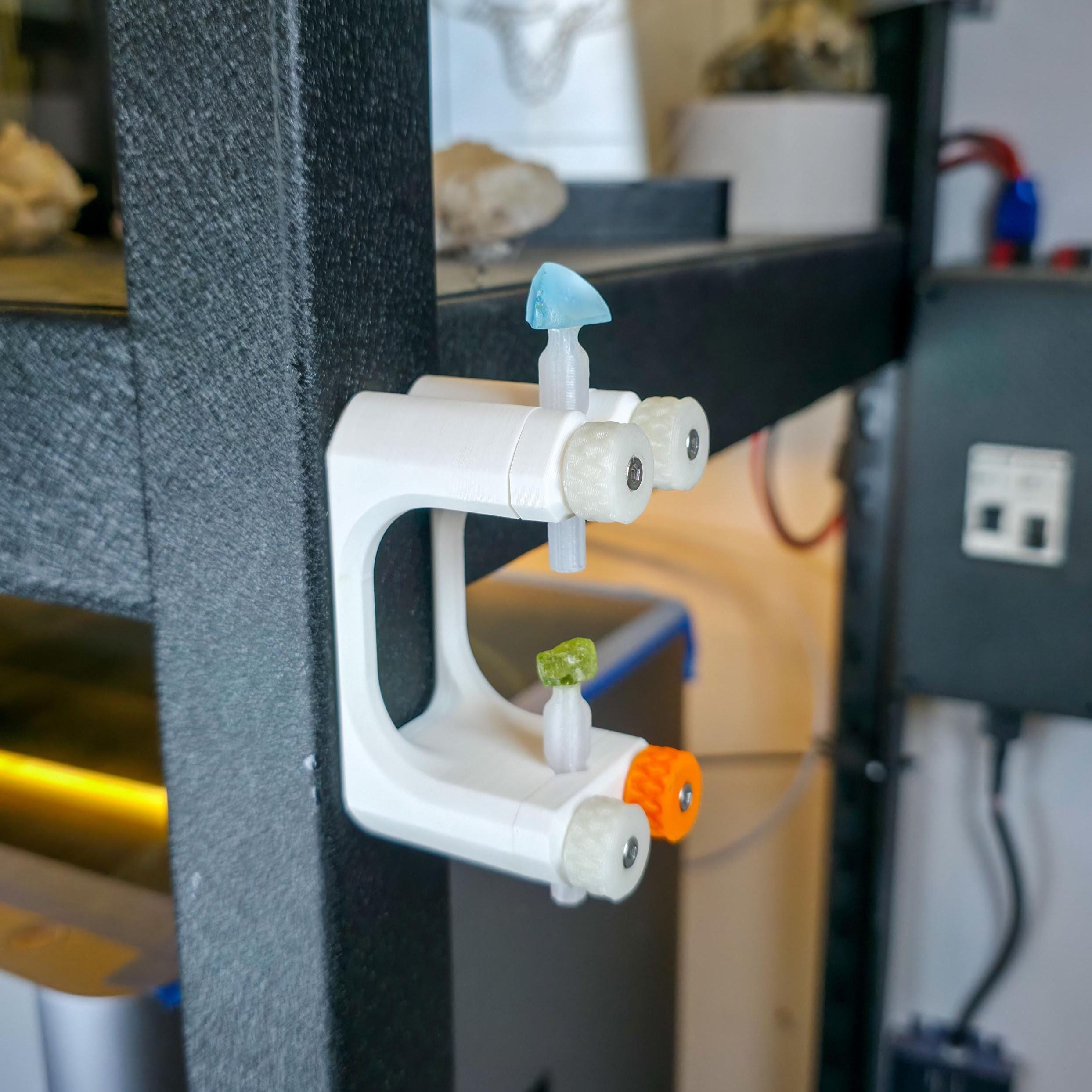 "A Gem Xfer fixture magnetically attached to a workshop frame, holding dop sticks with mounted blue and green gemstones. The setup keeps stones upright for curing or inspection, emphasizing practical studio integration and Aetherium’s modular magnetic storage system for precision lapidary work."