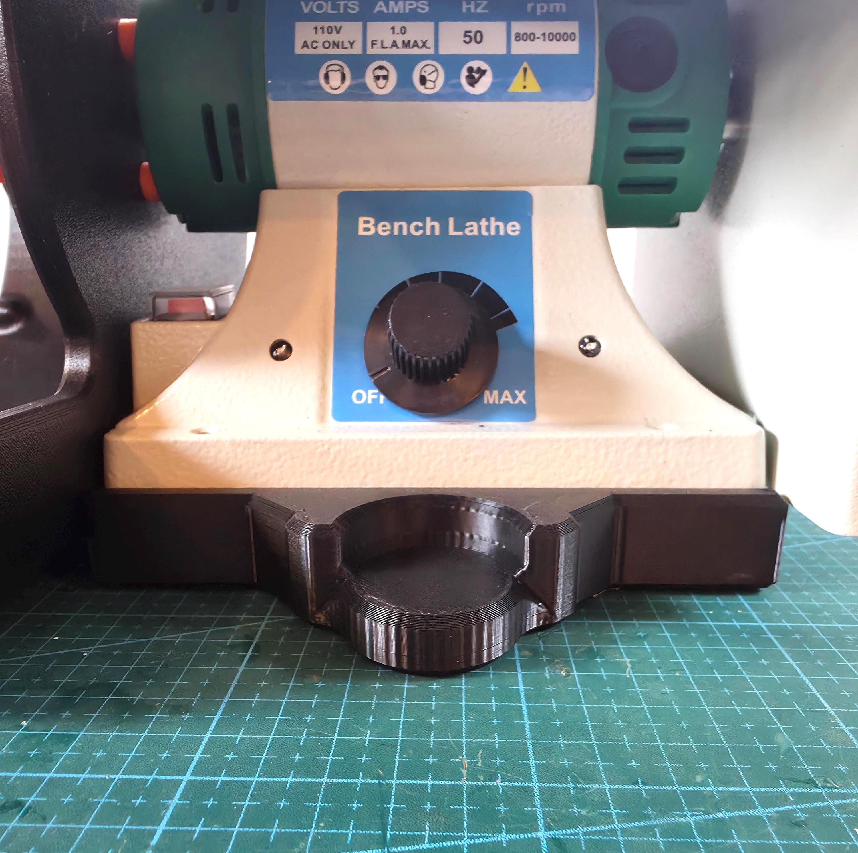 Front view of a 110V bench lathe motor mounted on the Aetherium Quick Clamp Adapter base, showing the form-fit circular clamp recess beneath the control dial. The adapter plate sits flush on a green cutting mat, designed for portable or fixed bench setups.