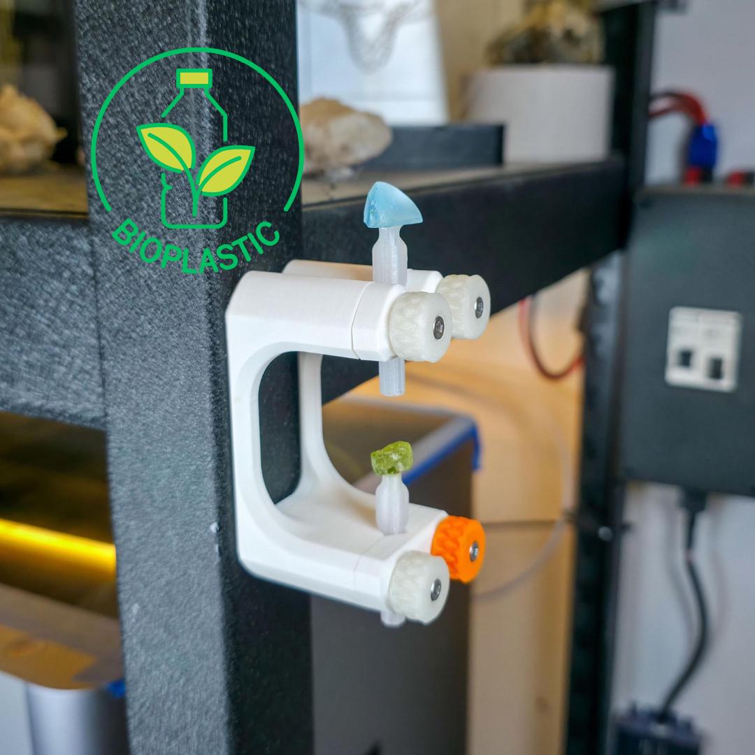 the gem Xfer is a 3d printed bioplastic lapidary jig that is hanging up on the wall using magnets. it's holding two dop sticks and has peridot and blue topaz attached with devcon 2 ton epoxy, and are curing in a verticle and level orientation.