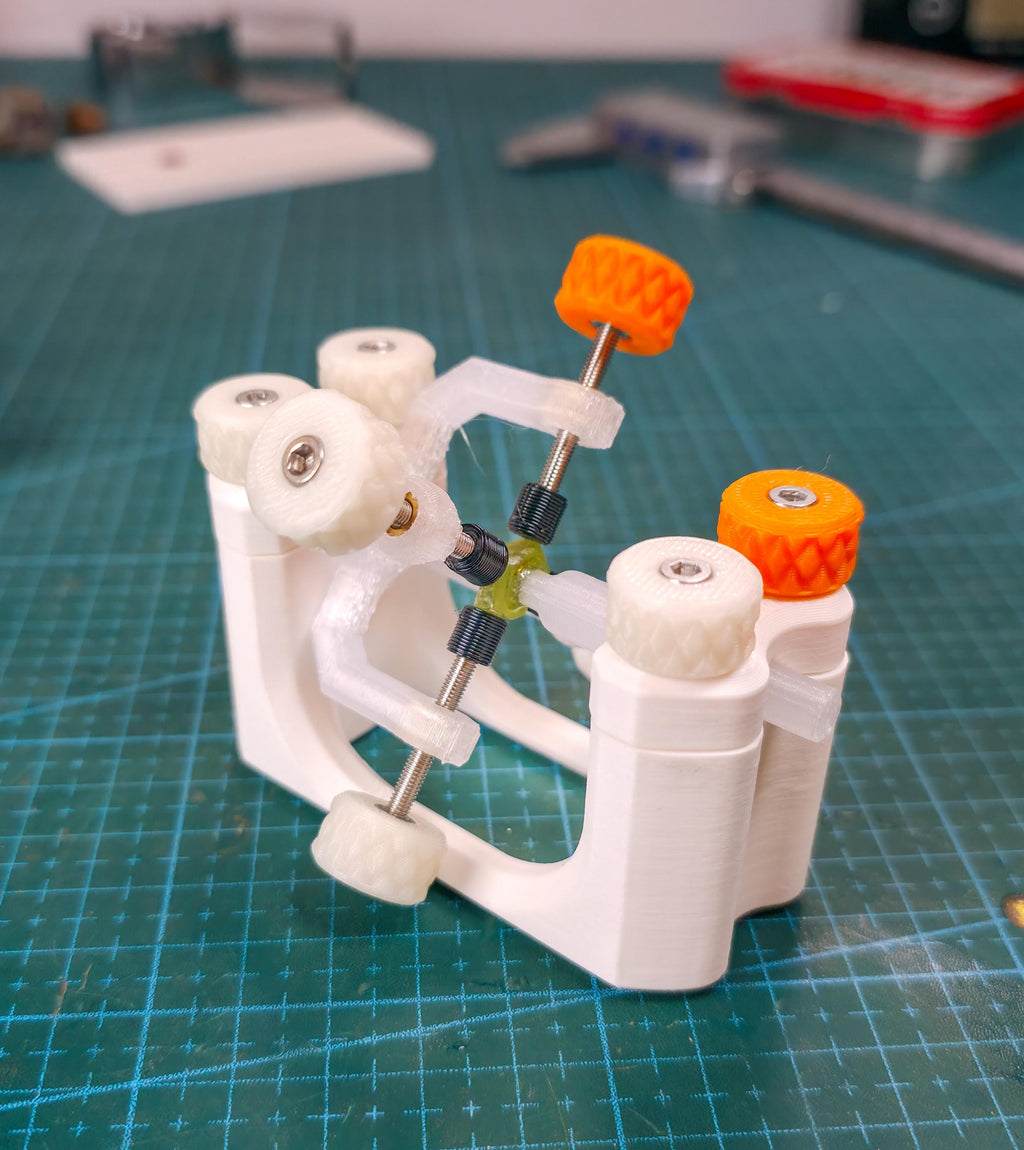 "The Gem Axis and Gem Xfer fixtures connected together on a cutting mat, showing the multi-axis adjustment system. Orange and white tension knobs and stainless-steel screws form a mechanical cross for angle control. The setup demonstrates the engineered adaptability of Aetherium’s lapidary tool system."