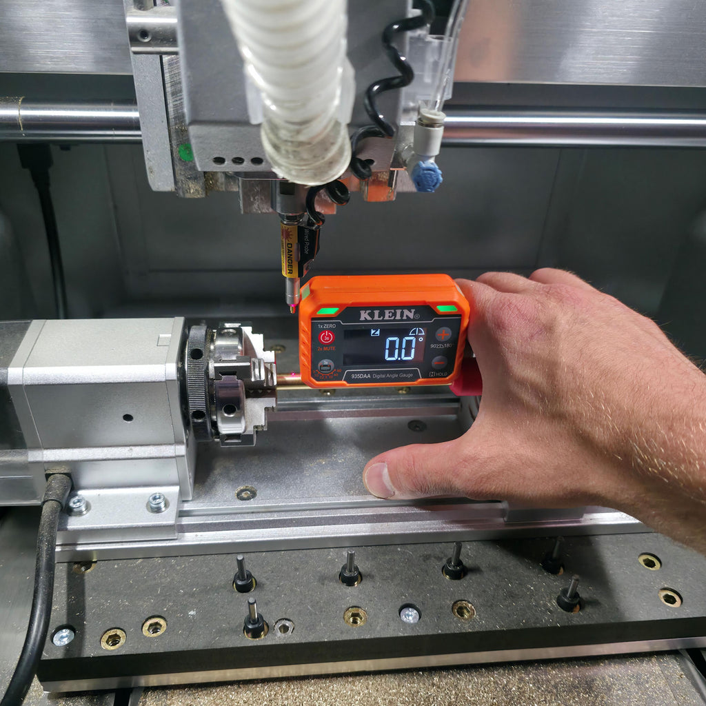 Operator’s hand using an orange Klein 935DAA digital angle gauge to check zero alignment on a Makera 4th-axis setup. A laser probe hovers above the centered brass workpiece inside the CNC enclosure.