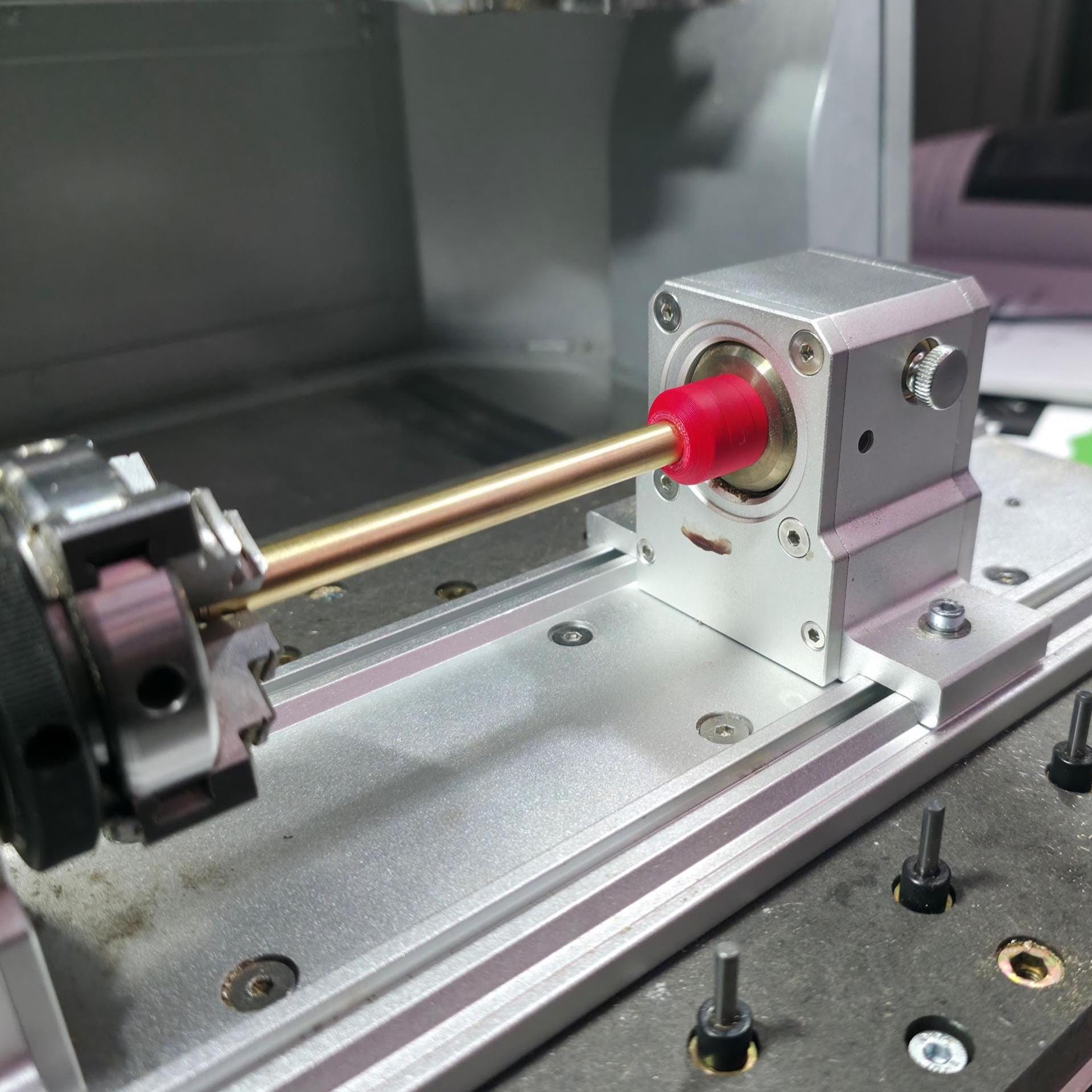 Makera 4th-axis rotary fixture mounted on an aluminum bed with a red centering jig installed on the pivot. Two knurled black tools rest nearby, surrounded by fixture bolts and machined dust.