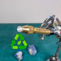 3D-printed clear polycarbonate dop stick inserted in brass faceting quill. Tip is rounded and layered for improved epoxy grip. Recycled material symbol shown on left. Tool is used in gemstone faceting, compatible with 6mm collets like Vevor systems. Set on a green cutting mat with rough stones in background