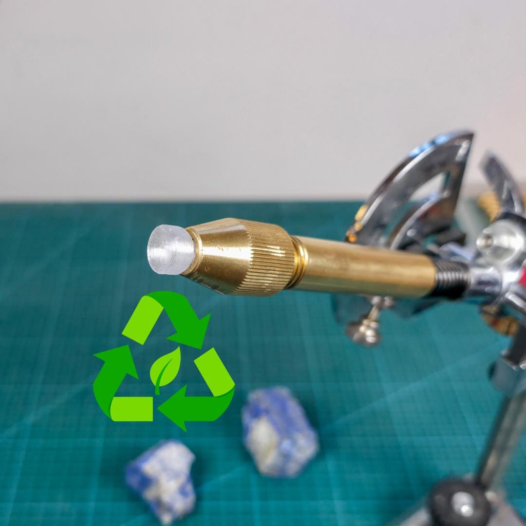 3D-printed clear polycarbonate dop stick inserted in brass faceting quill. Tip is rounded and layered for improved epoxy grip. Recycled material symbol shown on left. Tool is used in gemstone faceting, compatible with 6mm collets like Vevor systems. Set on a green cutting mat with rough stones in background
