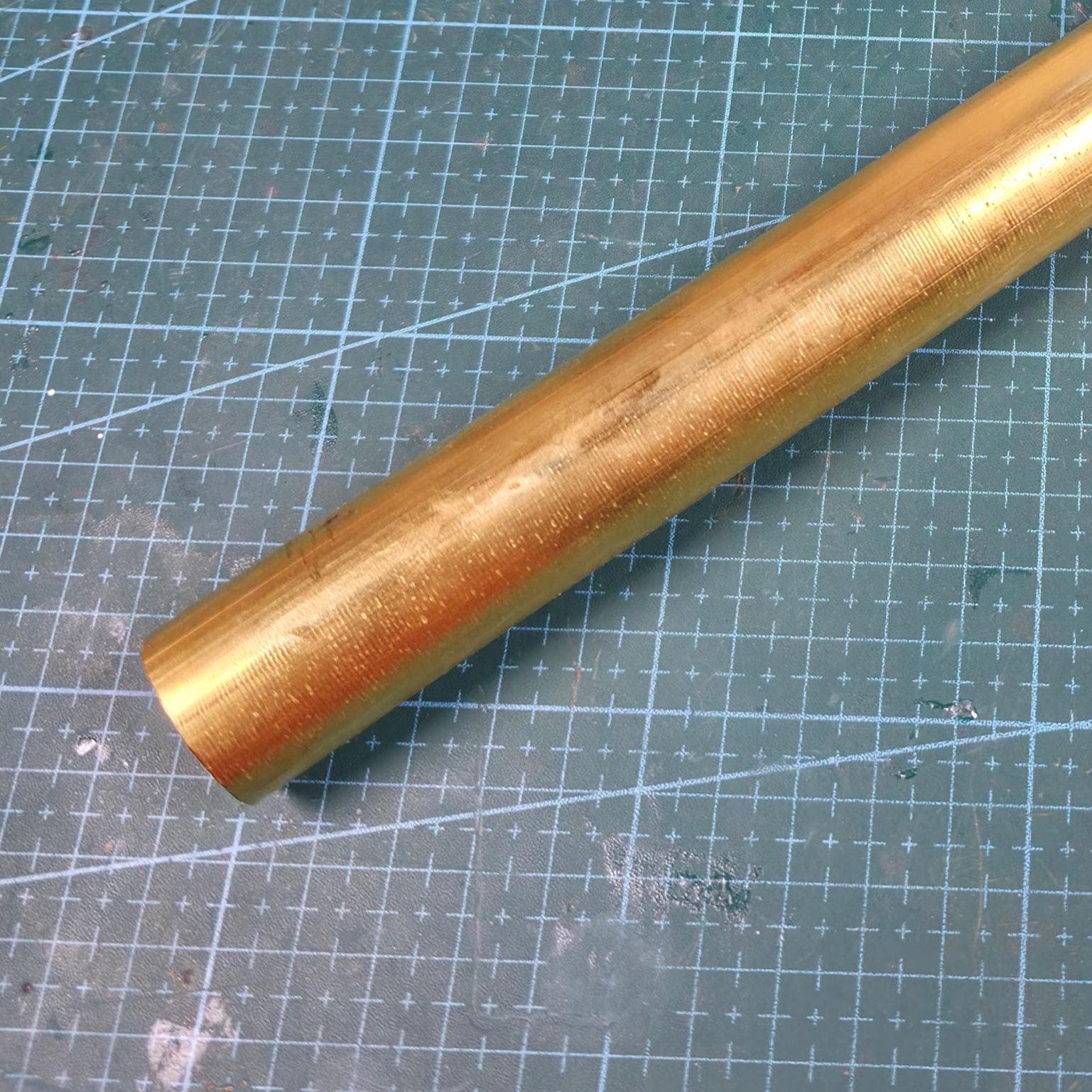 A cylindrical brass rod, angled across a green cutting mat. The rod has a golden hue and a slightly textured surface. The background features a grid pattern.