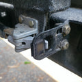 Black TPU swing gate isolator installed between a dual swing-out rear bumper latch and steel D-hook bracket. The flexible wedge compresses slightly to eliminate metal-on-metal contact, reduce vibration, and tighten latch closure on off-road tire carriers. Designed for overland vehicles like Toyota or Jeep, the isolator fits common rear bar setups and is shown fully seated in a rugged, trail-used hinge assembly.