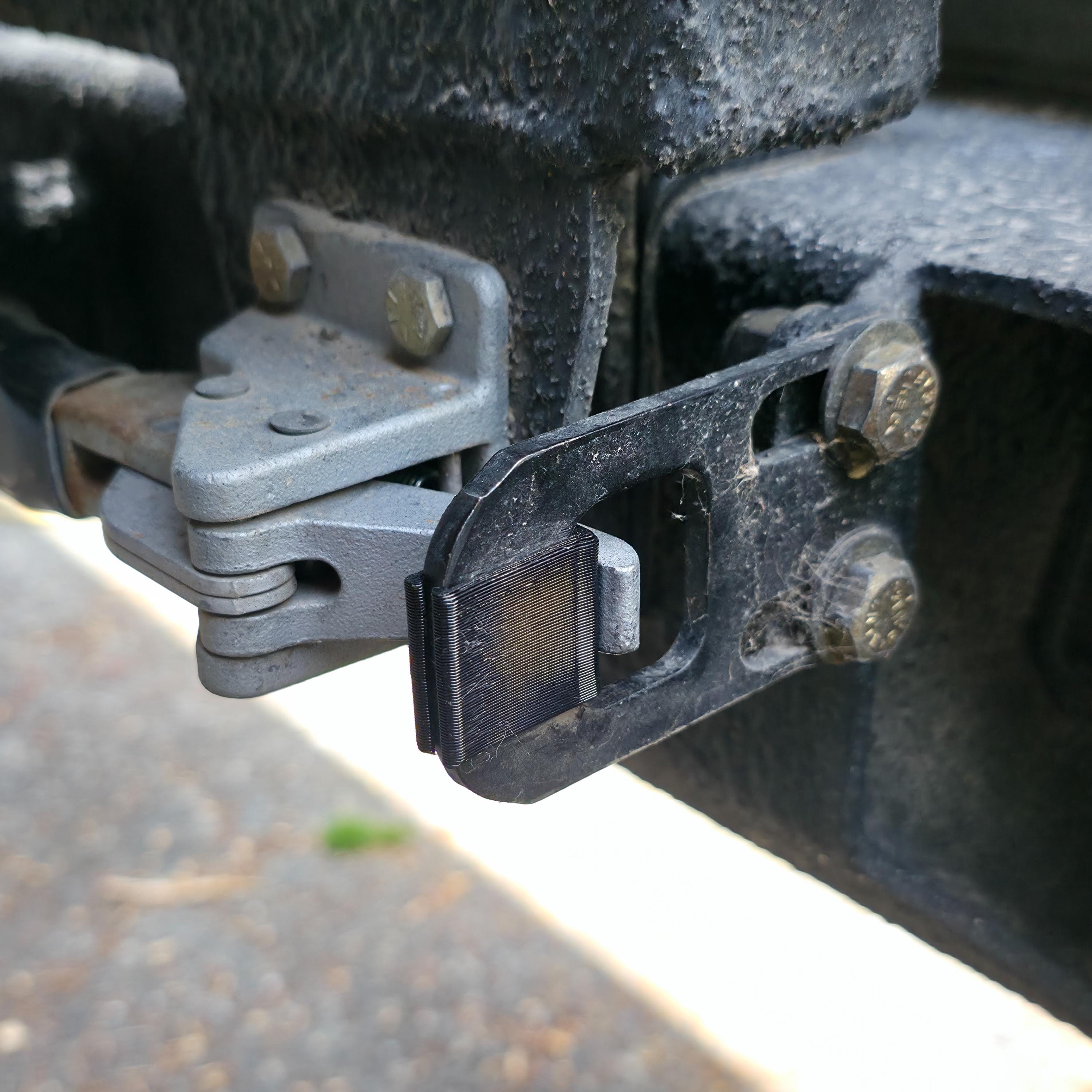 Black TPU swing gate isolator installed between a dual swing-out rear bumper latch and steel D-hook bracket. The flexible wedge compresses slightly to eliminate metal-on-metal contact, reduce vibration, and tighten latch closure on off-road tire carriers. Designed for overland vehicles like Toyota or Jeep, the isolator fits common rear bar setups and is shown fully seated in a rugged, trail-used hinge assembly.