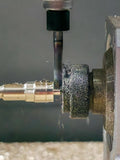 Close-up of a CNC mill cutting brass bar stock using a Makera 4th-axis attachment. A red alignment jig centers the workpiece as the spindle tool machines precise grooves, with metal chips visible on the surface.