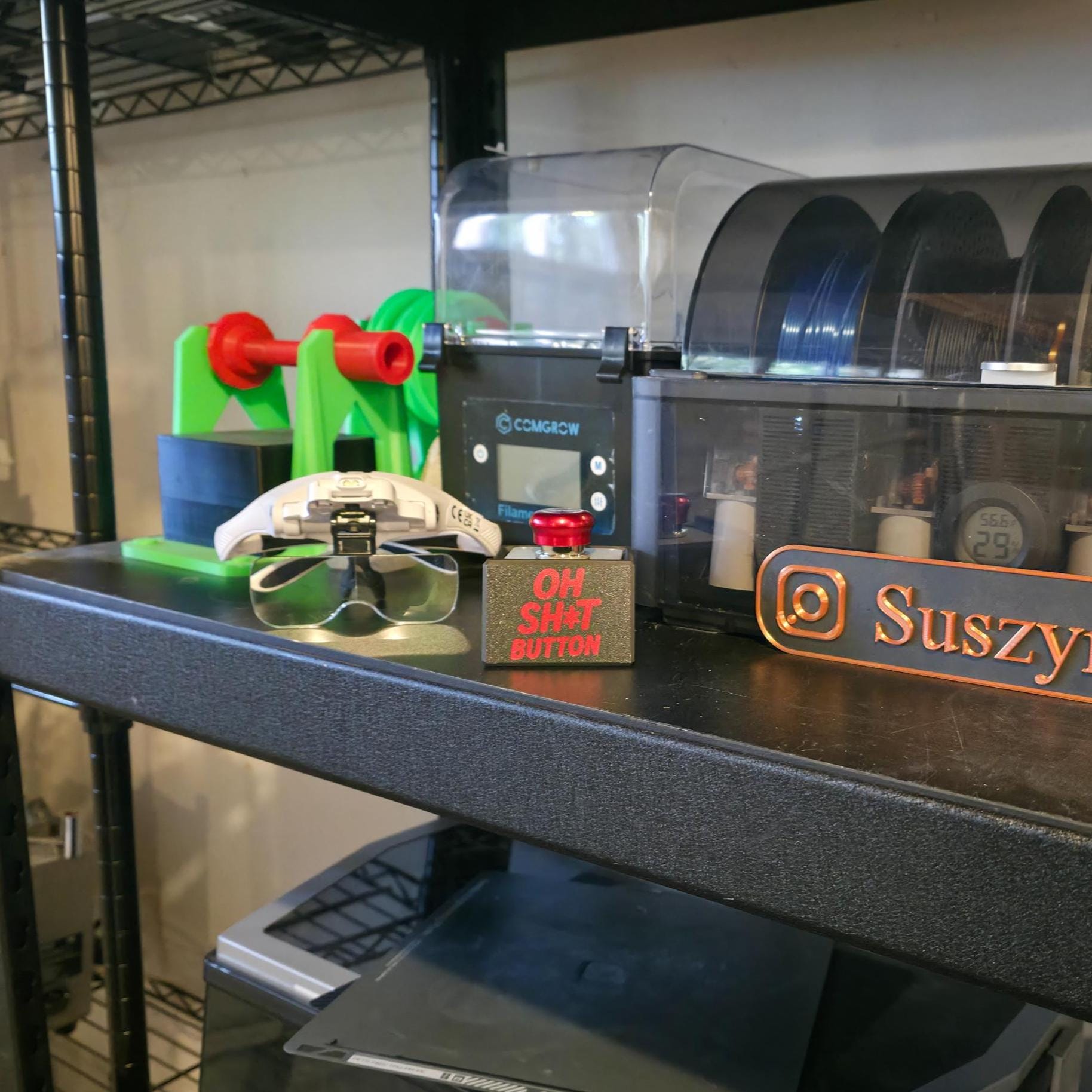 The “OH SH*T BUTTON” resting on a workshop shelf beside filament spools, green 3D printed parts, and safety glasses. The black metal box with a red metallic button adds a playful industrial touch to a modern maker workspace or CNC setup.