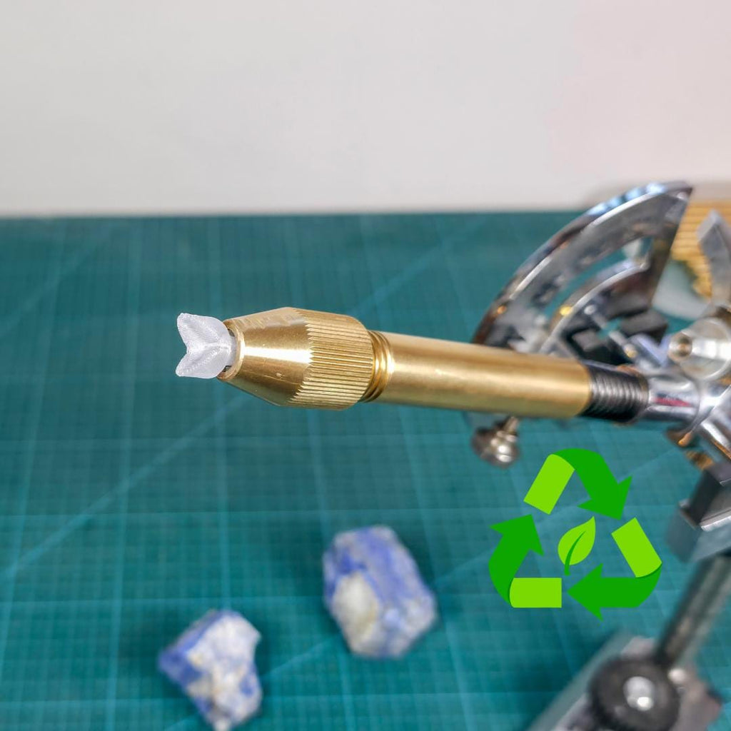 3D-printed clear polycarbonate dop stick inserted in brass faceting quill. Tip is shaped to hold a pavilion and layered for improved epoxy grip for cutting the stone face. Recycled material symbol shown on left. Tool is used in gemstone faceting, compatible with 6mm collets like Vevor systems. Set on a green cutting mat with rough stones in background.