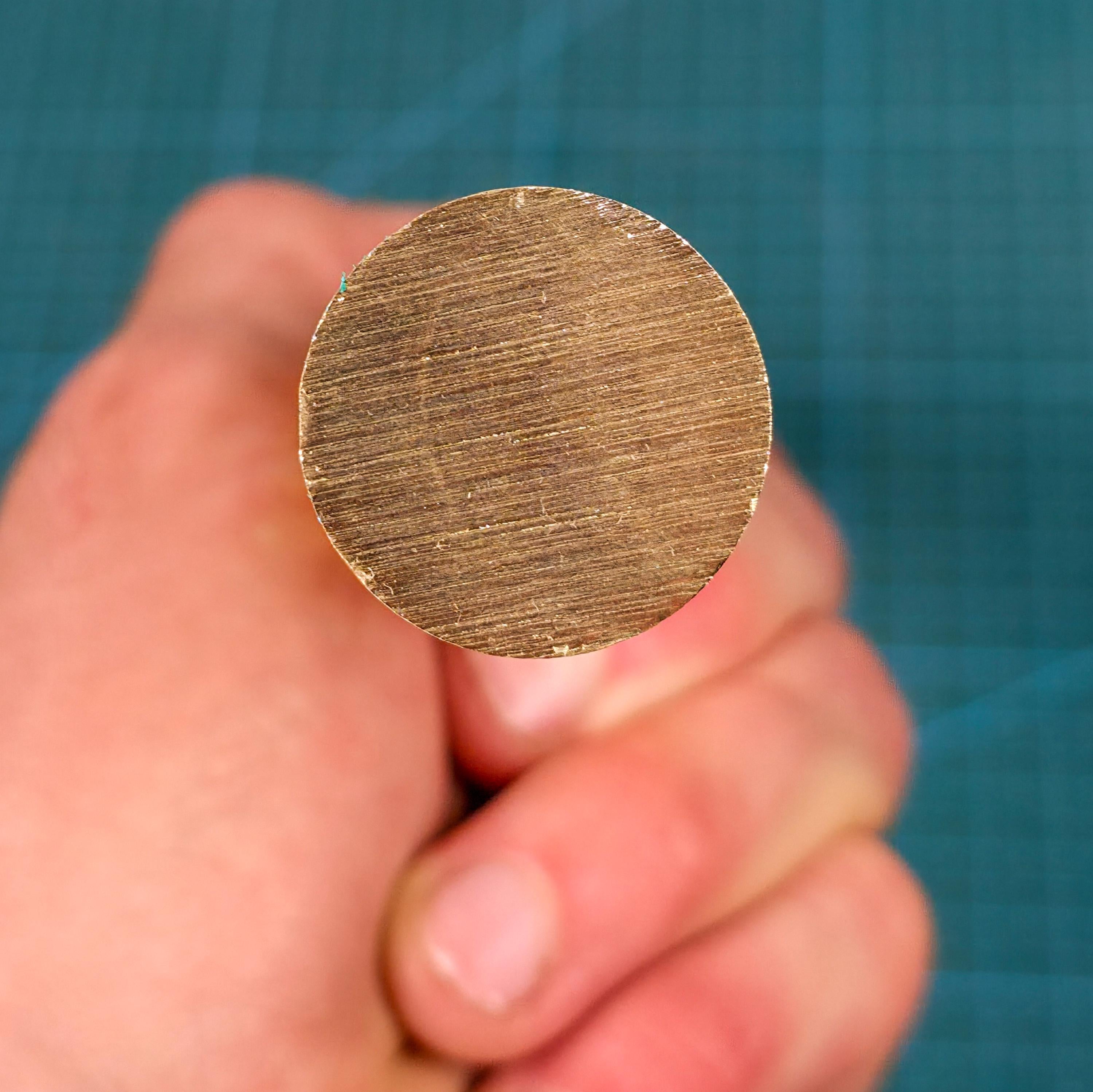 Close-up photograph of the end of a brass round stock rod held between fingers for scale. The circular face shows uniform material grain and clean cut edges. The rod diameter is 1 inch, equivalent to 25.4 millimeters, highlighting real-world scale and machinable solid brass stock for lathe and CNC use.