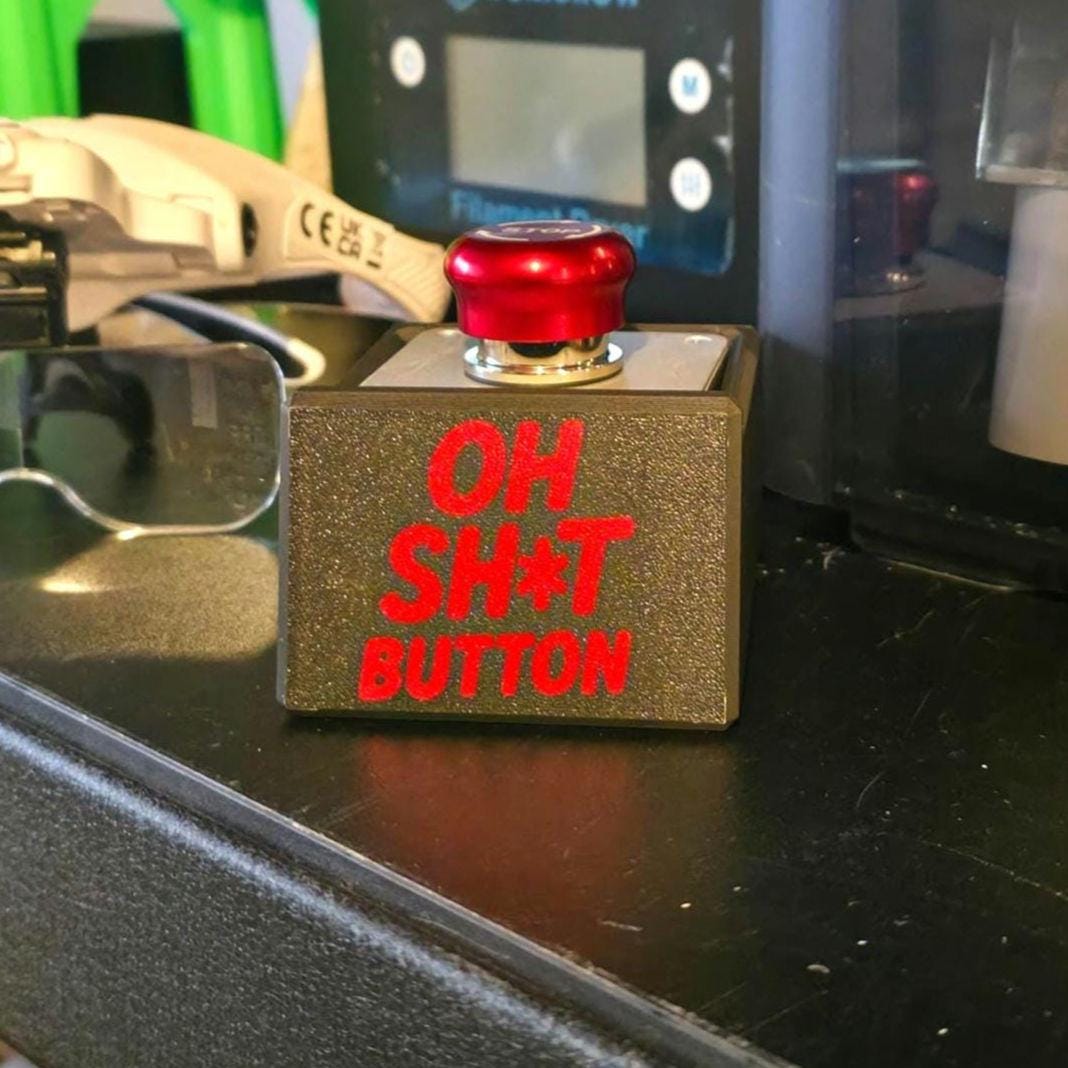 Close-up of a handmade emergency-style red push button labeled “OH SH*T BUTTON” in bold red text on a matte black housing. The button sits on a black workshop bench surrounded by maker tools and CNC equipment, used as a humorous accessory for 3D printers, CNC machines, or creative workstations.