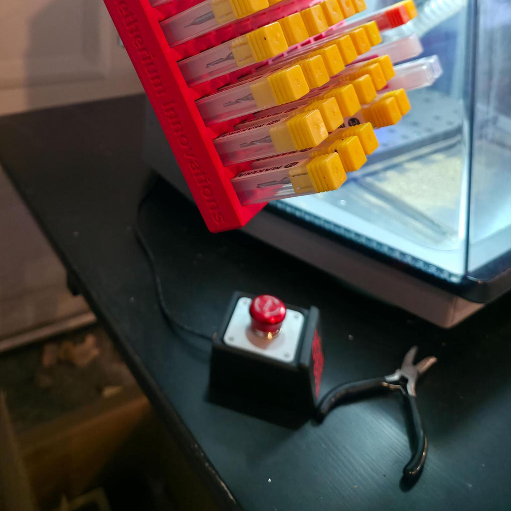 The “OH SH*T BUTTON” connected near a Carvera Air CNC machine, shown with tools and accessories on a dark bench. The button’s bold red text and metallic top stand out under warm workshop lighting, designed as a fun emergency-style control for makers.