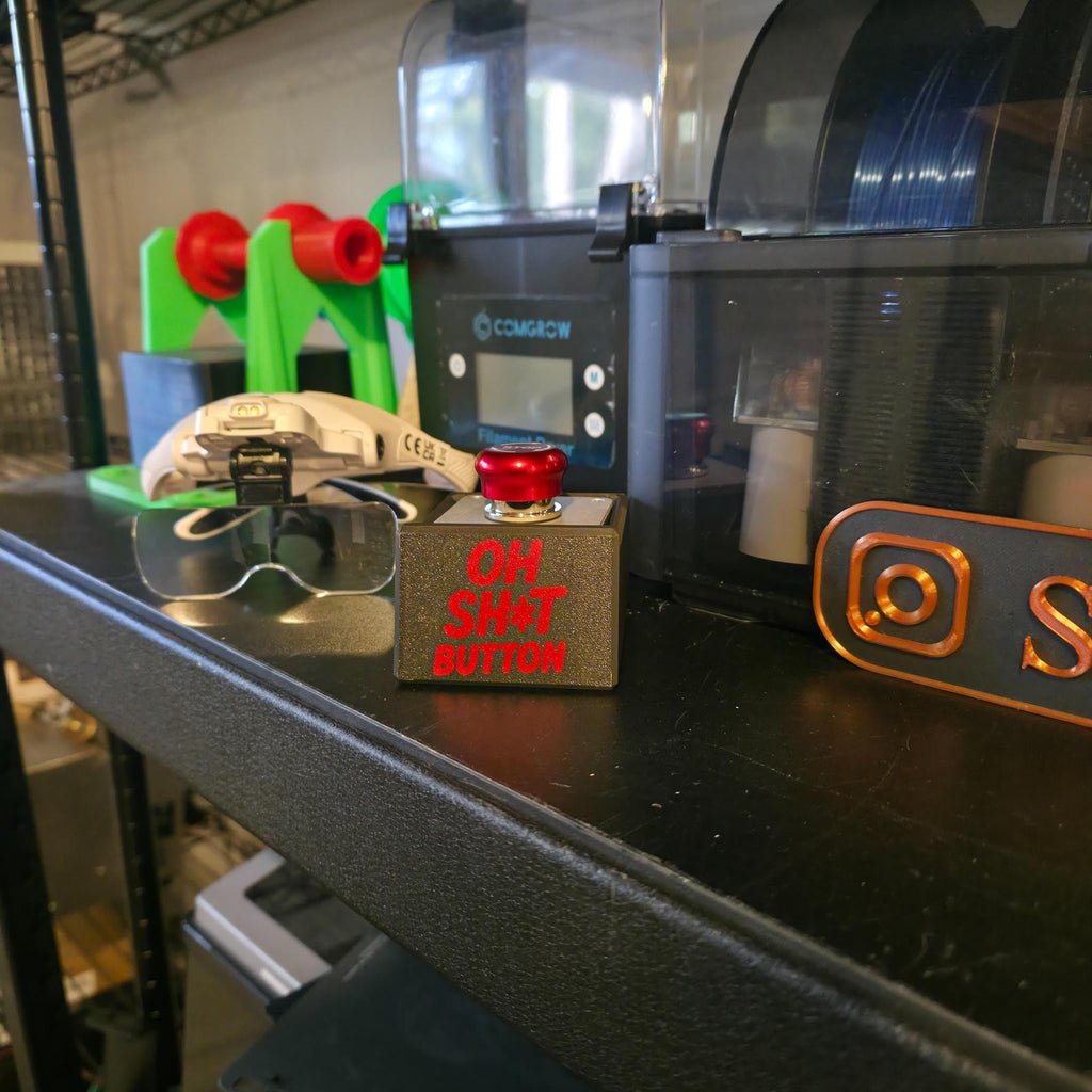 Side view of the “OH SH*T BUTTON” beside 3D printing accessories and maker tools on a black shelf. The red engraved lettering contrasts sharply against the black surface, showcasing a handmade, industrial aesthetic.