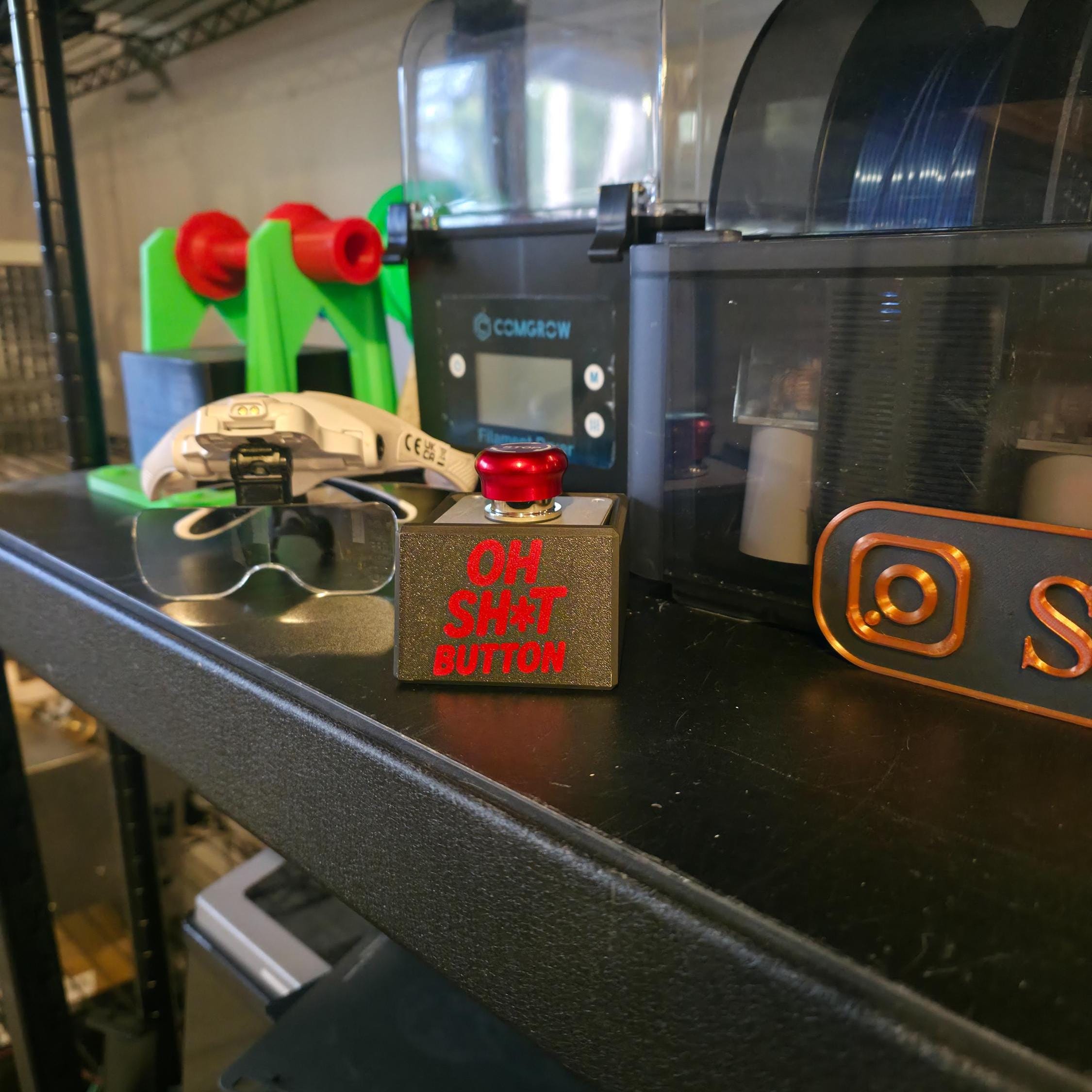 Side view of the “OH SH*T BUTTON” beside 3D printing accessories and maker tools on a black shelf. The red engraved lettering contrasts sharply against the black surface, showcasing a handmade, industrial aesthetic.