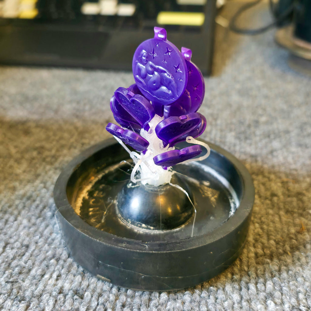 Purple 3D-printed wax medallion masters mounted on a central sprue for lost-wax casting, shown attached to a rubber base mold cap. Each medallion features a detailed embossed figure of a reclining woman surrounded by stars, arranged in a circular cluster for jewelry production. The wax sprue system highlights casting preparation for making multiple identical pendants. perfect for resin, hand card for 3d printed projects.