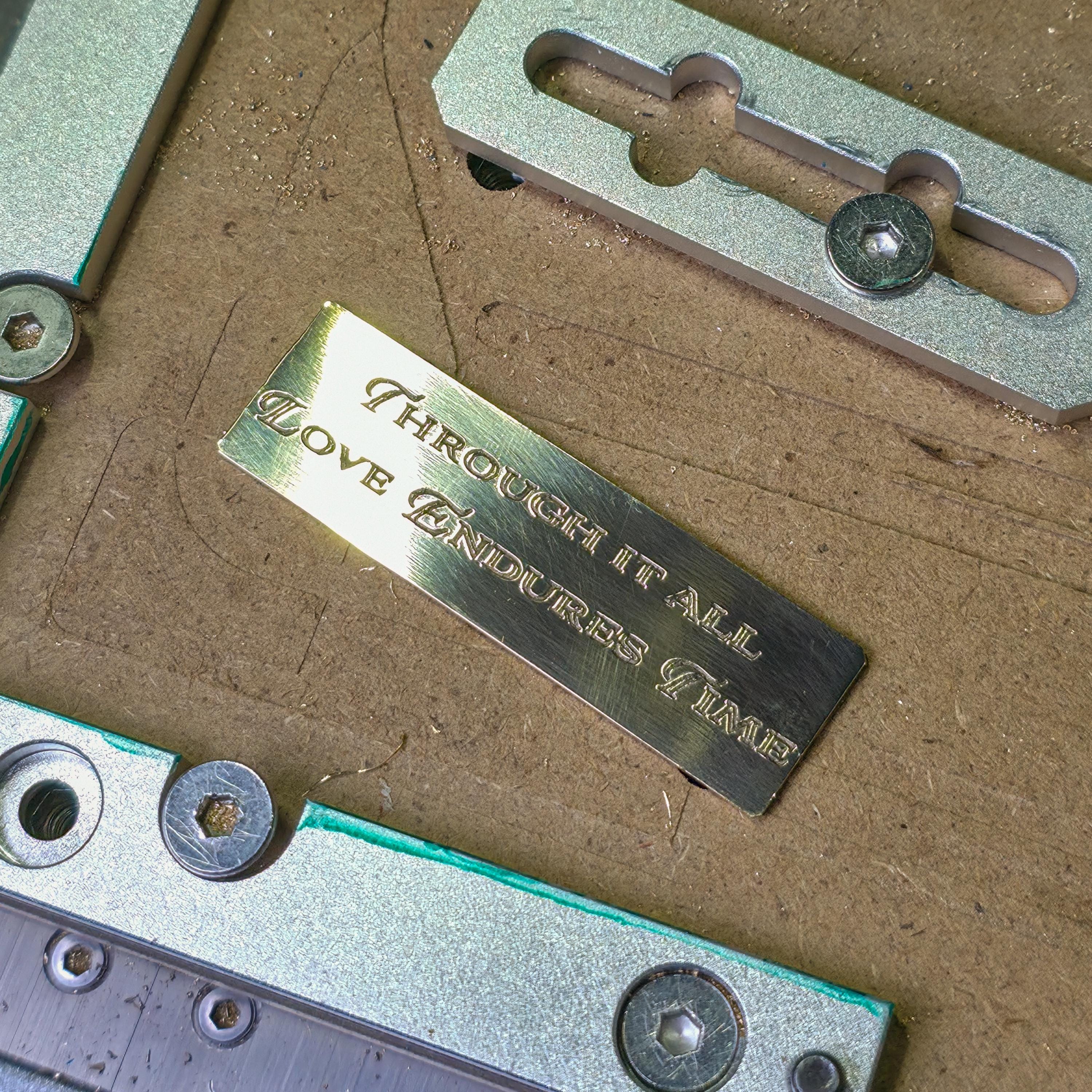 Brass plaque engraved with the phrase “Through it all, love endures time,” held in a metal fixture on a workshop table. The close-up reveals crisp lettering, polished gold tones, and fine machining detail.