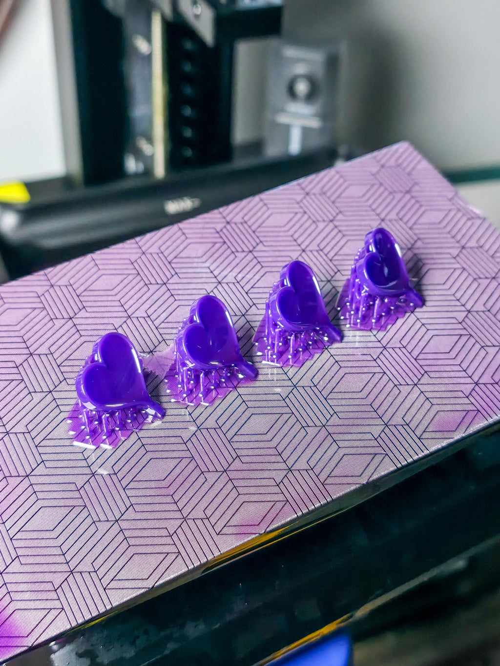 Four purple heart pendants freshly 3D printed in resin, shown attached to a patterned build plate inside a workshop. The vibrant color and precise supports highlight early-stage jewelry prototyping.