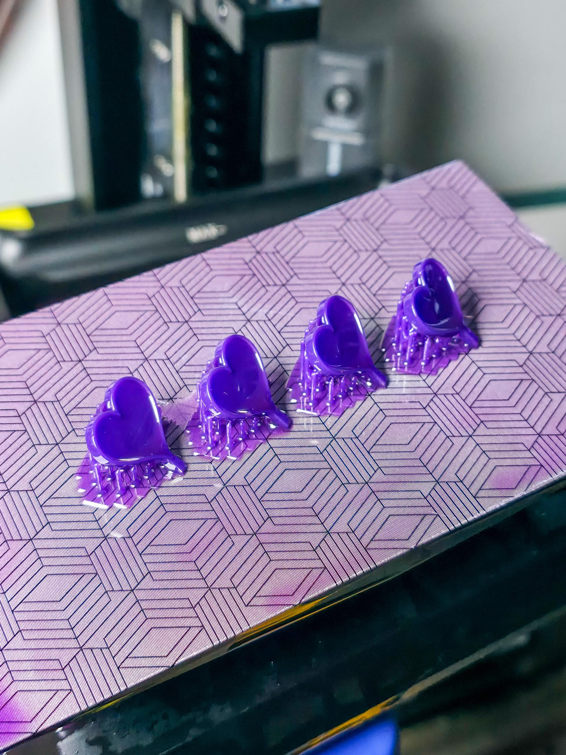 Four purple heart pendants freshly 3D printed in resin, shown attached to a patterned build plate inside a workshop. The vibrant color and precise supports highlight early-stage jewelry prototyping.