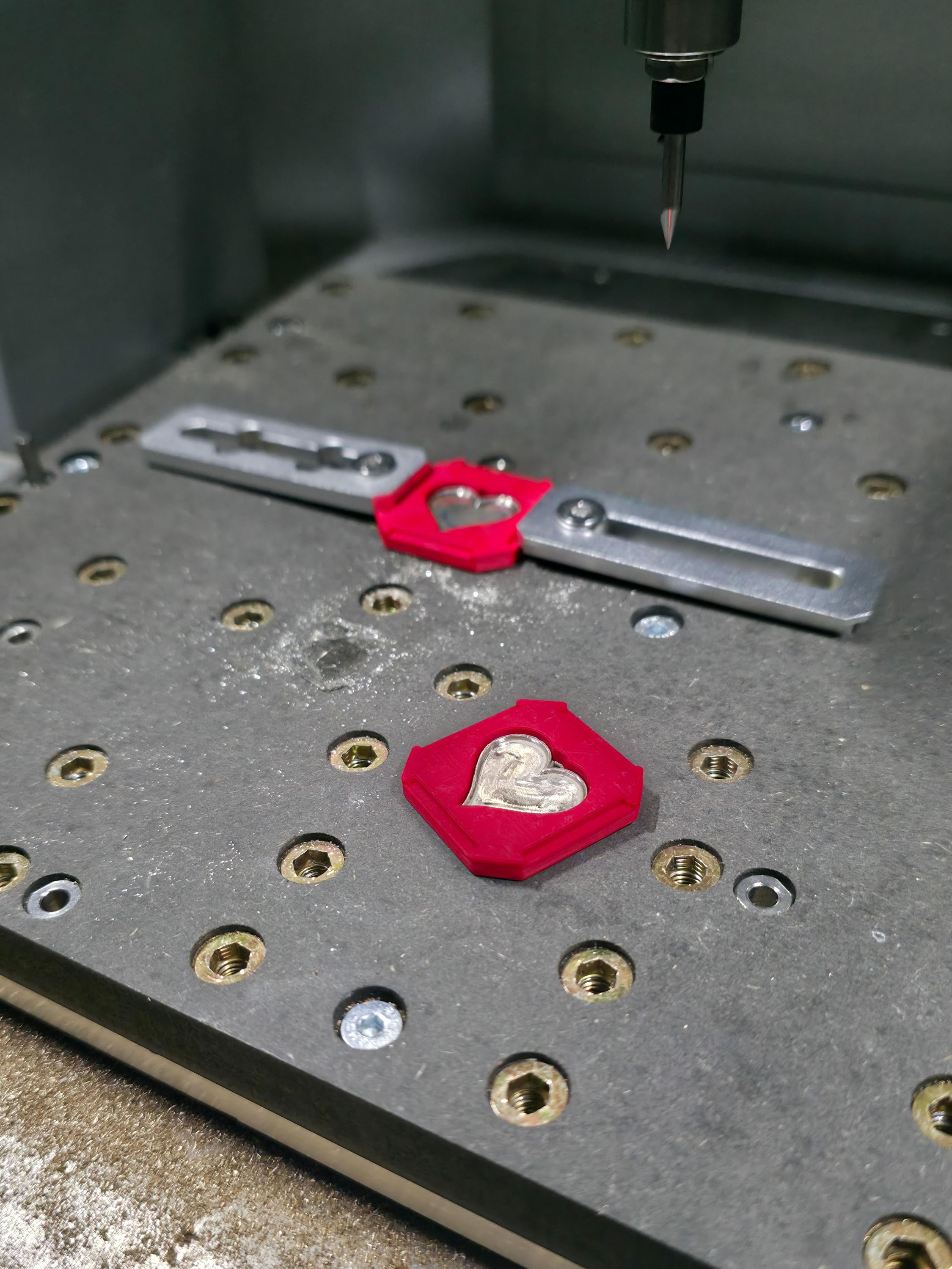 Two metallic heart pendants set into red fixtures during CNC engraving. The machine bed and cutting bit visible above convey the precision manufacturing process behind Aetherium’s fine metal jewelry.