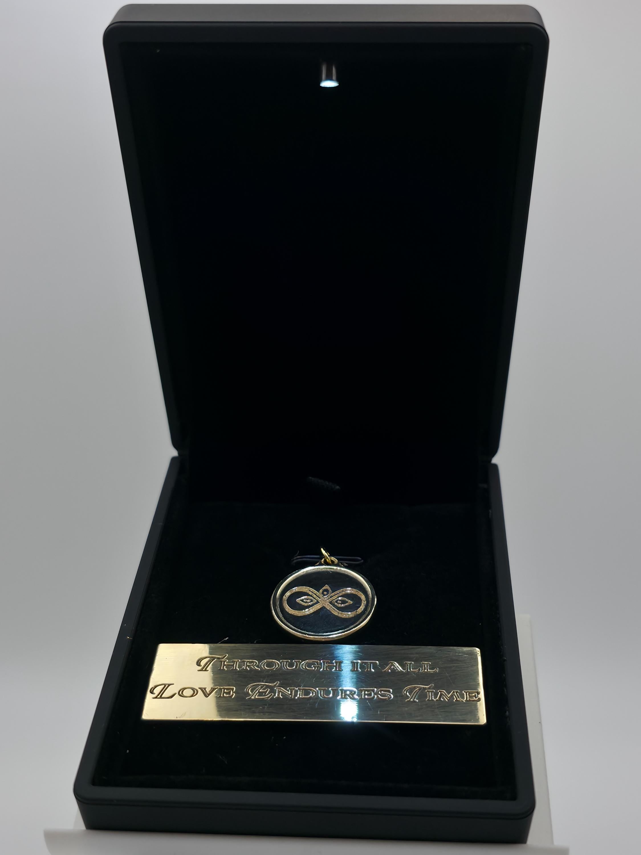 Infinity pendant in brass shown under LED-lit luxury jewelry box. The built-in light highlights the engraved infinity and lotus design while paired with a polished brass plaque. Presentation emphasizes premium quality and gift appeal for couples, anniversaries, and milestone celebrations.
