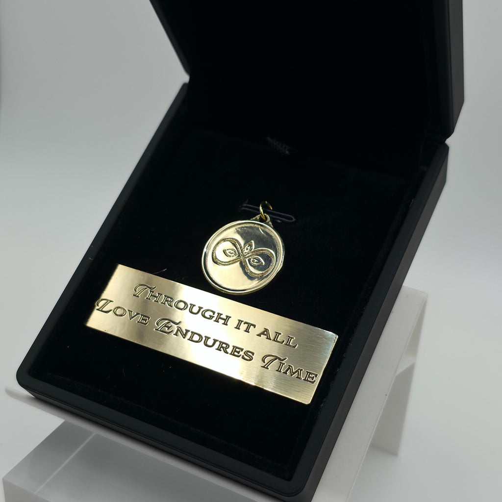 Angled view of the Aetherium Infinity Pendant in solid brass, resting in black velvet with matching brass plaque inscribed “Through it all, love endures time.” Hand-polished finish and engraved design catch the light, emphasizing artisan craftsmanship and timeless symbolism in a gift-ready presentation.