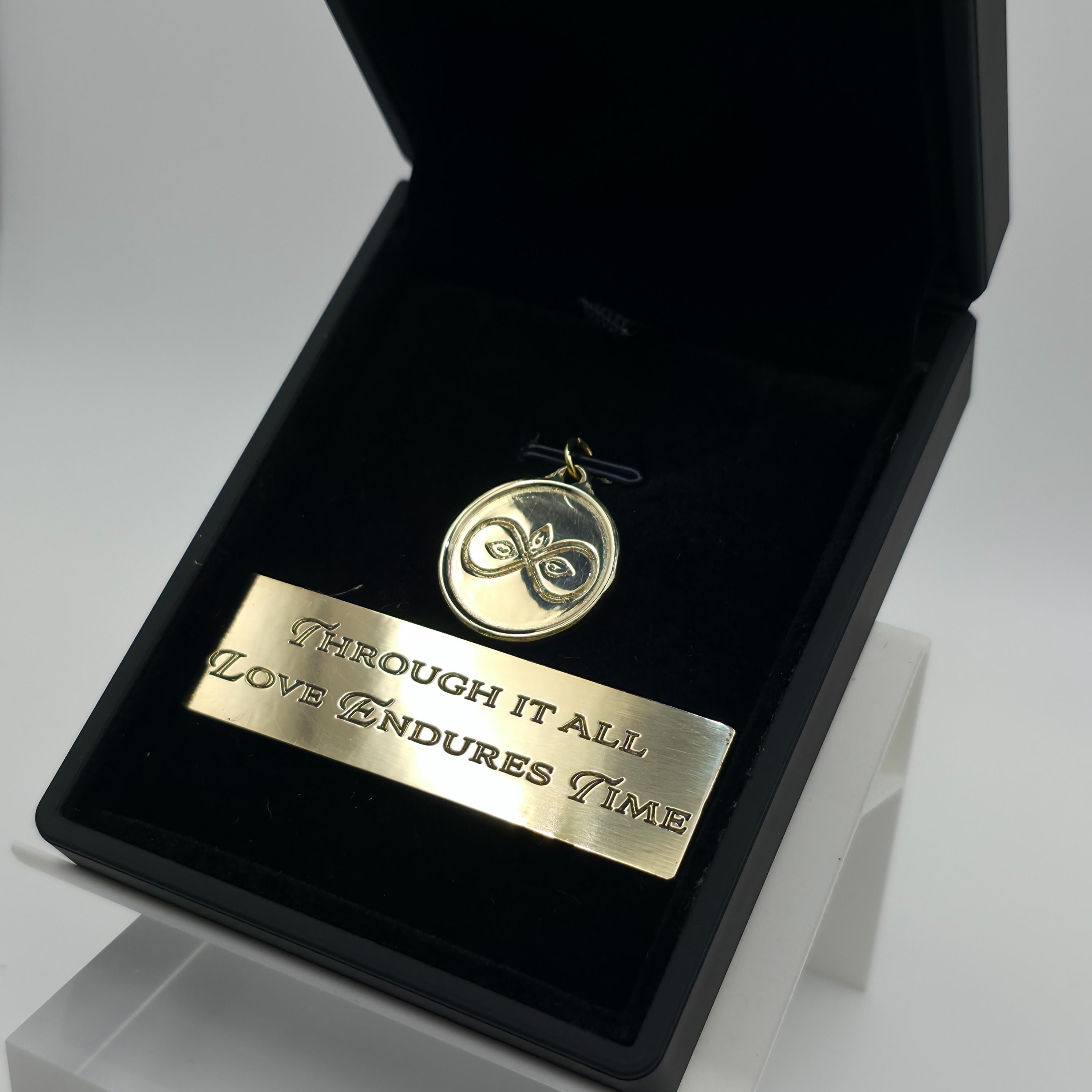 Angled view of the Aetherium Infinity Pendant in solid brass, resting in black velvet with matching brass plaque inscribed “Through it all, love endures time.” Hand-polished finish and engraved design catch the light, emphasizing artisan craftsmanship and timeless symbolism in a gift-ready presentation.