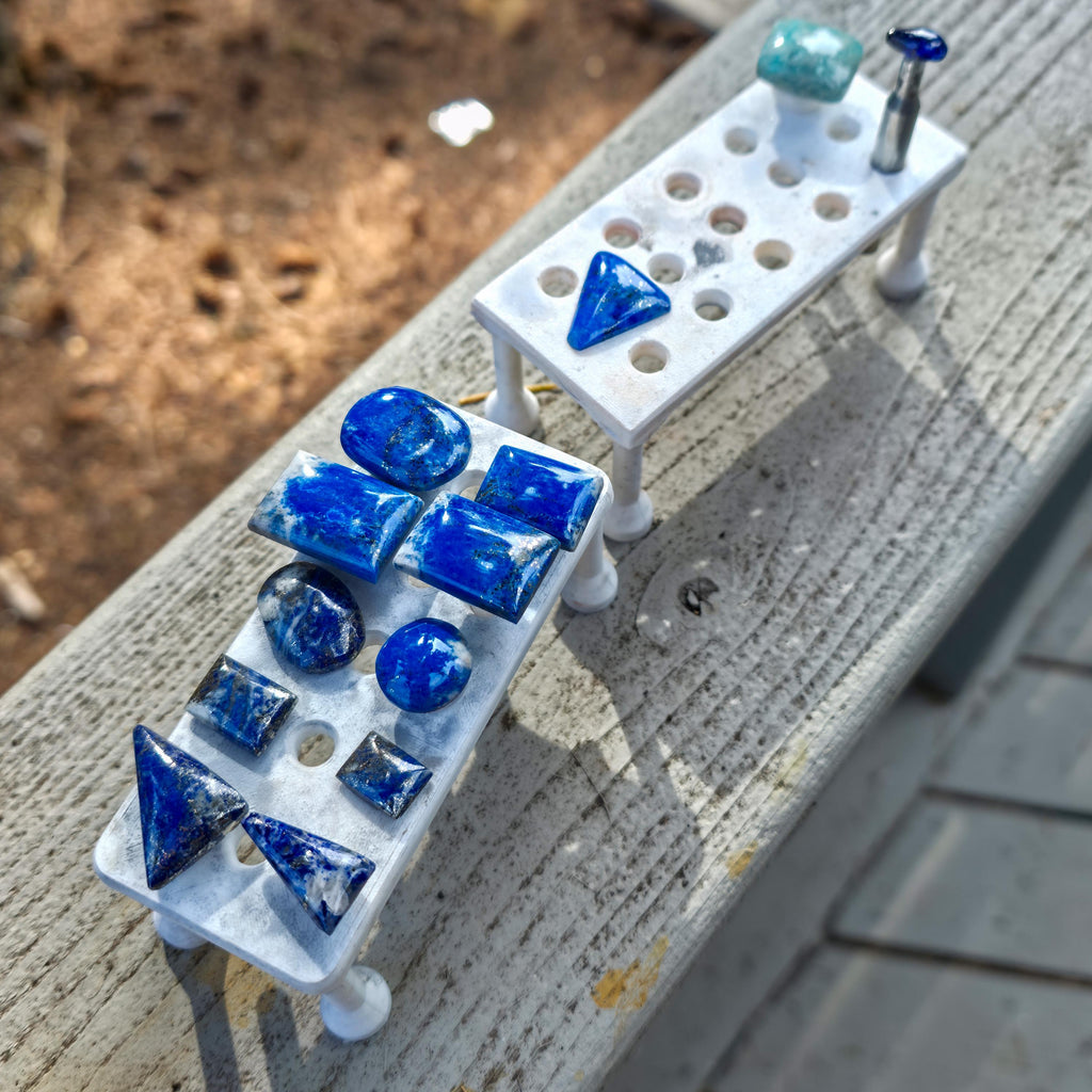 Two 3D-printed magnetic dop stick trays placed on a wooden rail outdoors, holding polished blue lapis lazuli cabochons and dop sticks, demonstrating stable staging, visibility, and organization of stones during lapidary cutting and polishing processes.