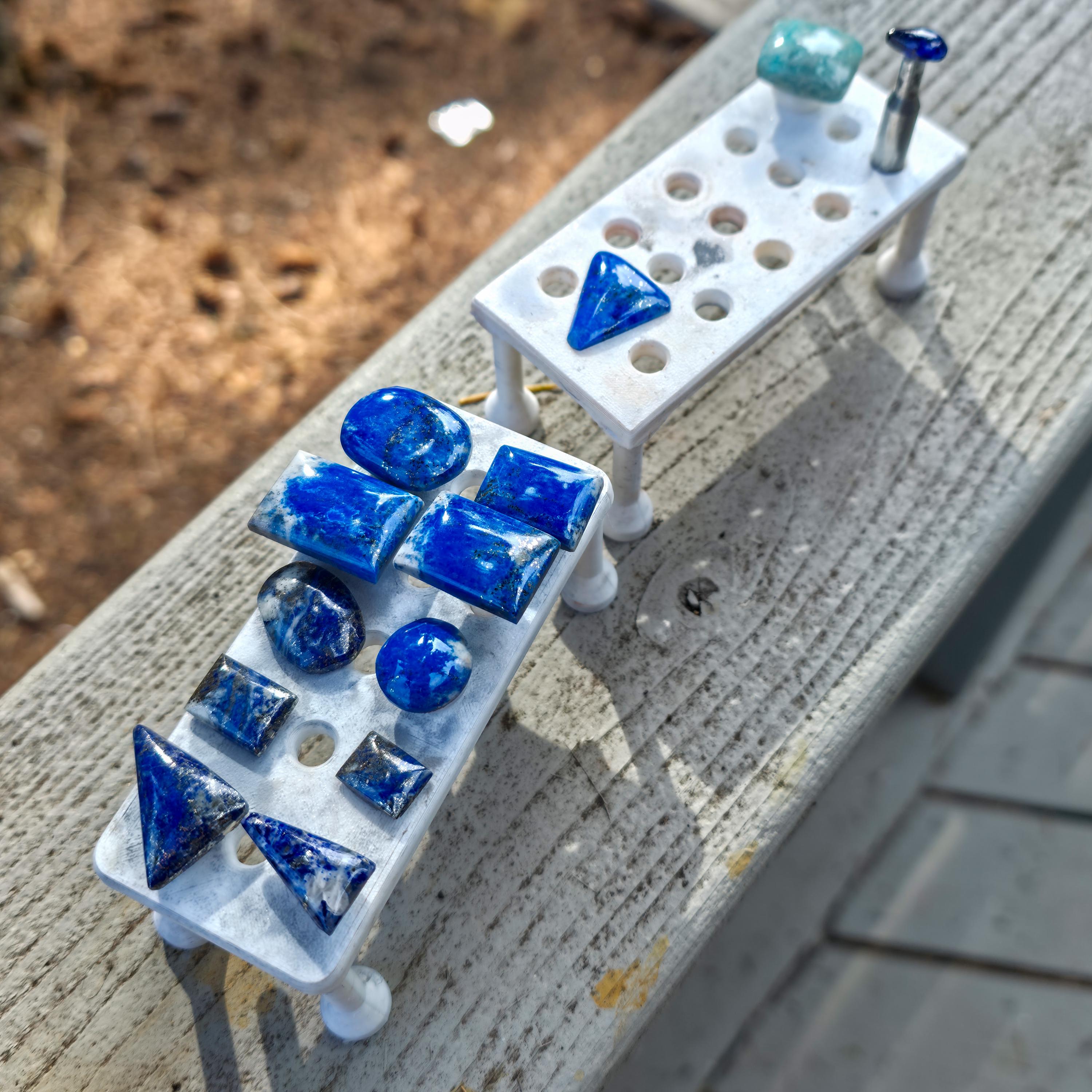 Two 3D-printed magnetic dop stick trays placed on a wooden rail outdoors, holding polished blue lapis lazuli cabochons and dop sticks, demonstrating stable staging, visibility, and organization of stones during lapidary cutting and polishing processes.