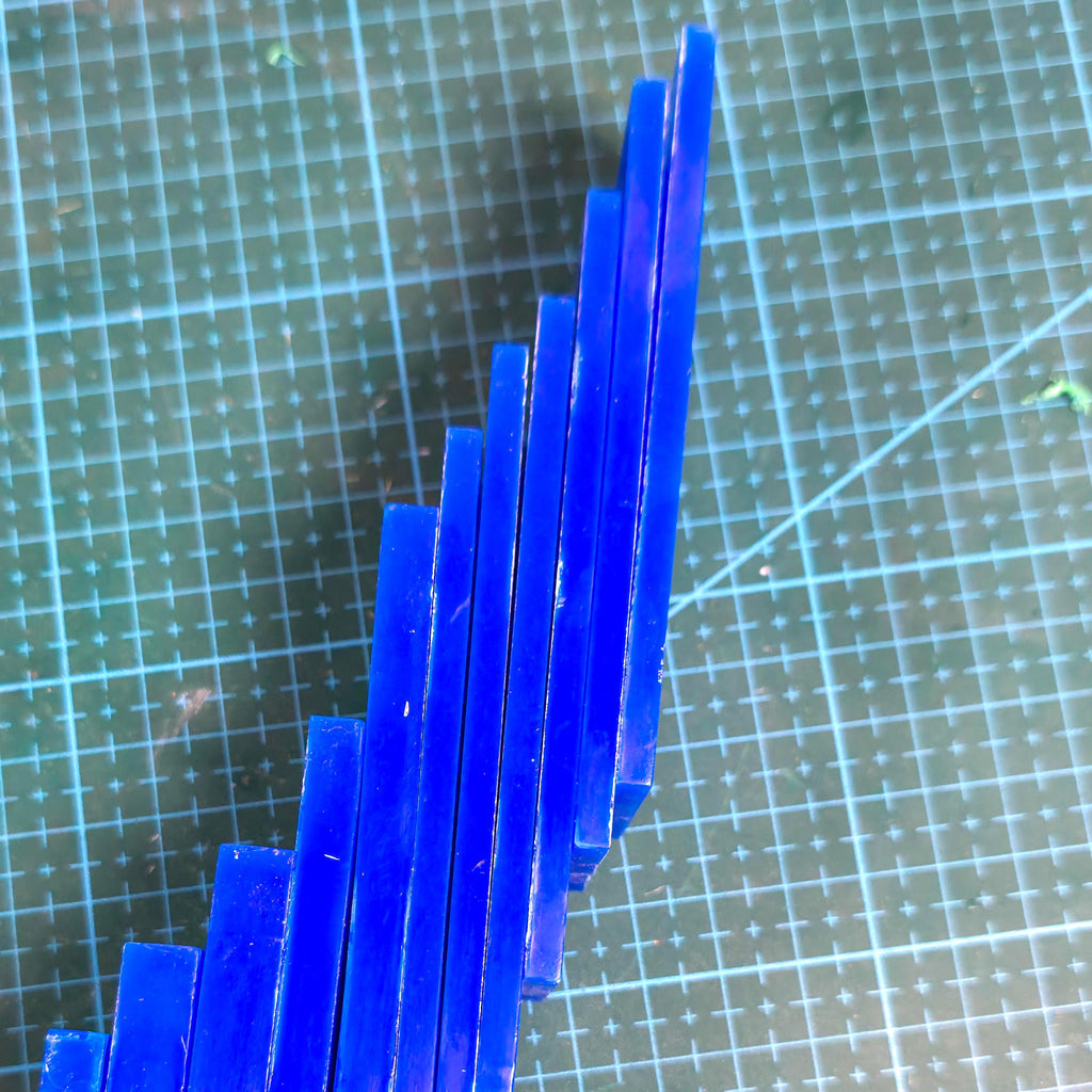 Top view of blue carving wax blocks stacked vertically in increasing thickness on a grid cutting mat. Each block shows smooth, even surfaces for use in lost-wax jewelry casting and model carving.