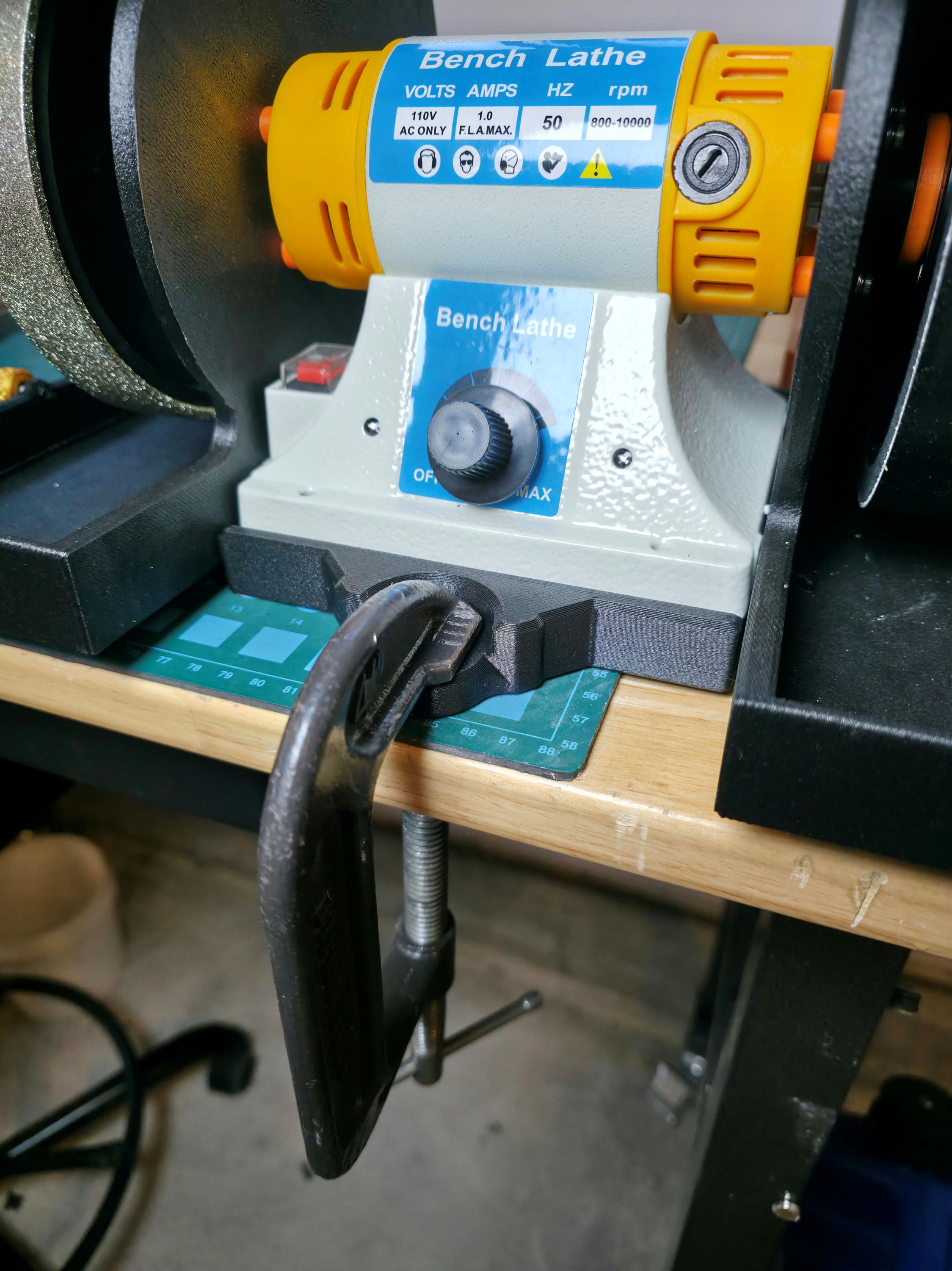 Bench lathe motor secured to a wooden workbench using the Aetherium Quick Clamp Adapter base with a metal C-clamp for portable mounting and vibration reduction.