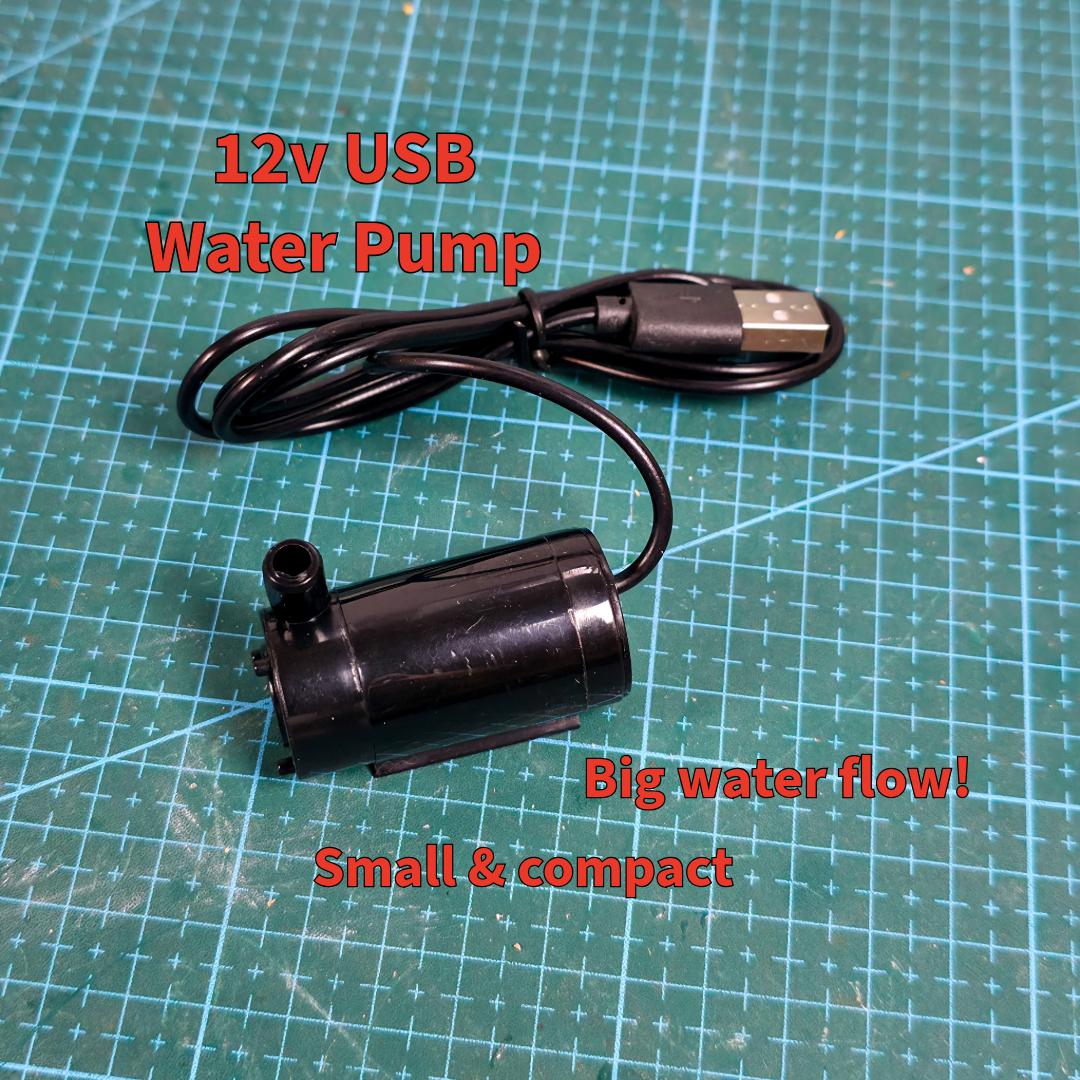 Close-up of a black USB-powered 12V water pump resting on a cutting mat. Text notes its high flow rate and compact size. This pump powers the MakerSpace Edition’s active water cooling system, suitable for portable use and small workspace setups.