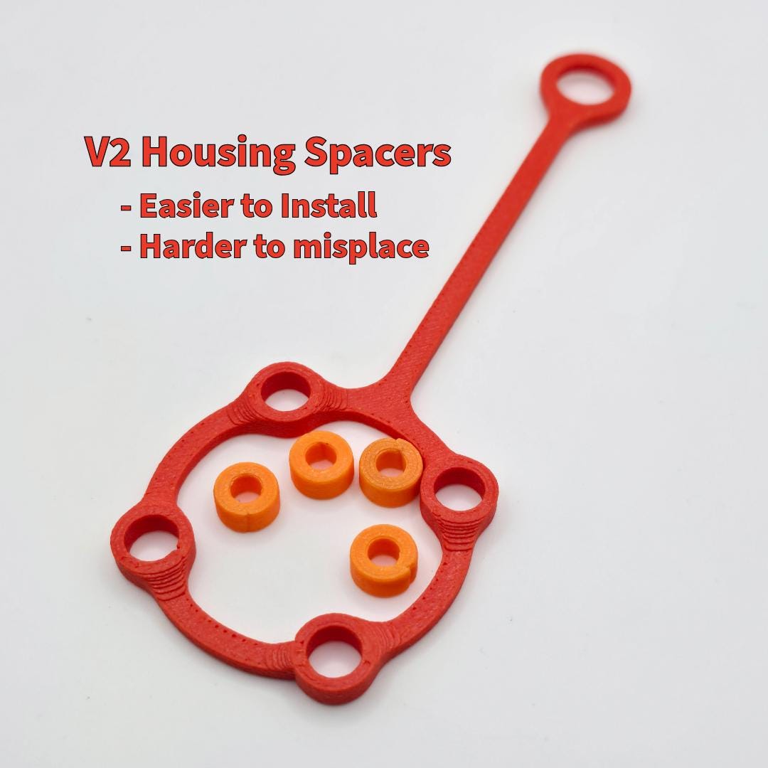 Image of red and orange polymer housing spacers linked together on a single mounting strap. Caption emphasizes improved installation and reduced risk of loss. These spacers maintain consistent alignment and spacing within the machine housing for accurate wheel fitment and easy servicing.