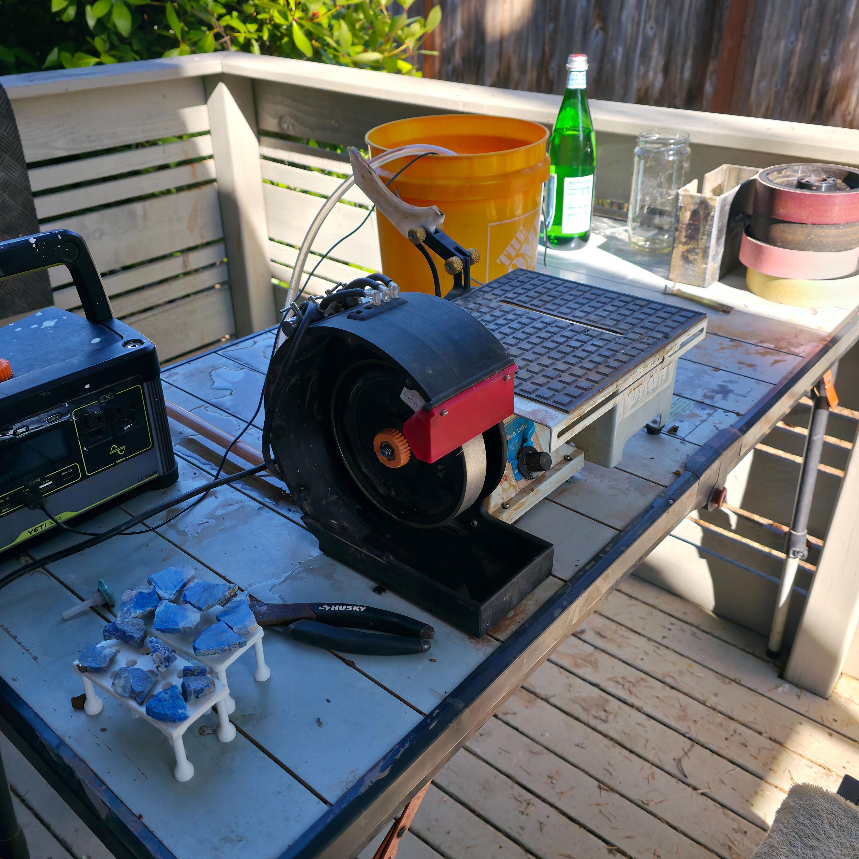 Wide shot of the MakerSpace Edition system in use on an outdoor workbench. The grinder, saw table, and magnetic stone holders are visible with supporting tools and a portable power station. Demonstrates real-world lapidary operation in a workshop or field environment.