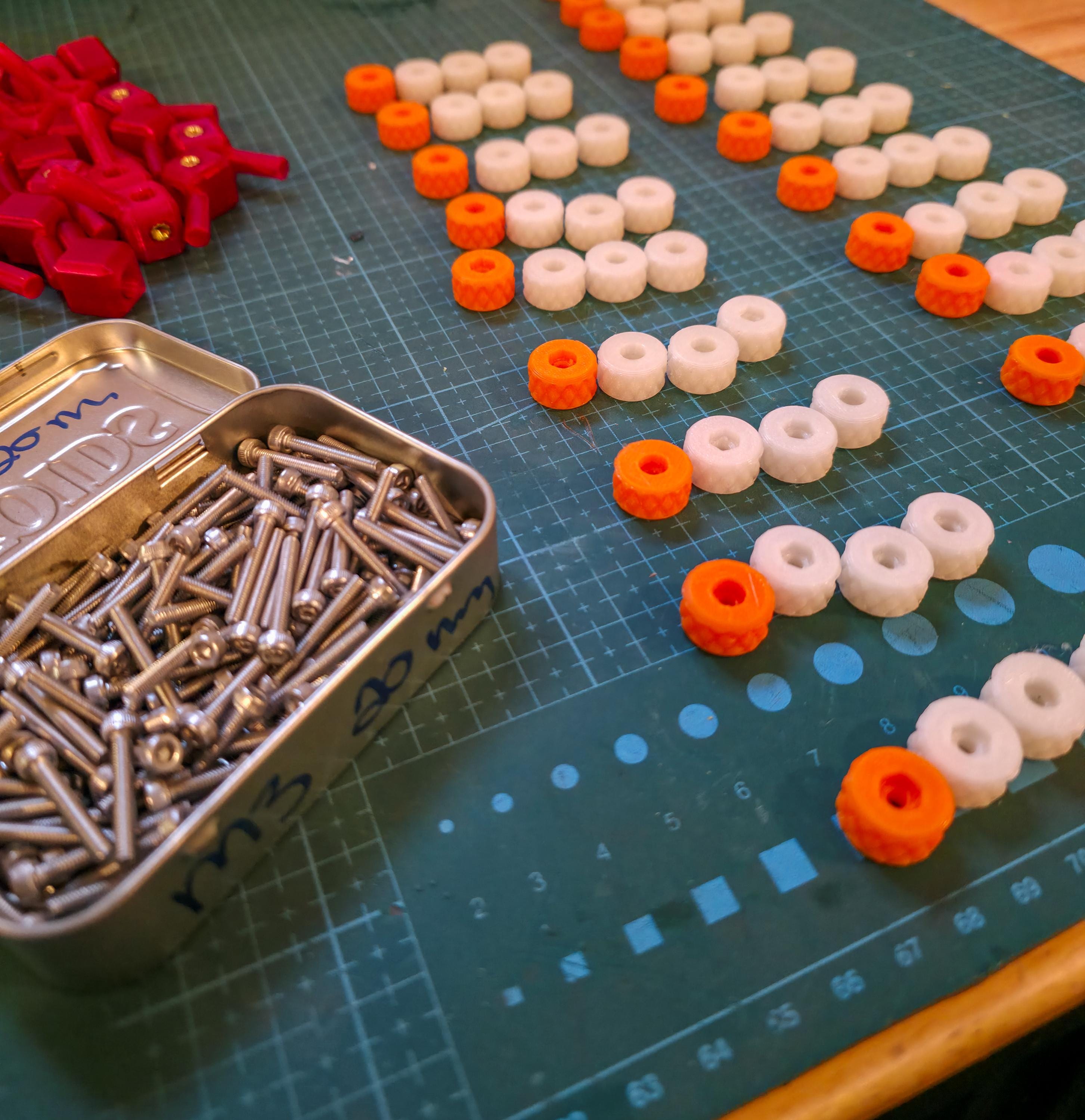 Knurled knobs and machined screws arranged in organized batches on a cutting mat, with a tin of M3 hardware. Different knob colors indicate variants. Scene highlights the modular fastening system for the Gem-Xfer jig, showing what builders need when assembling the digital STL design for gemstone epoxy curing.