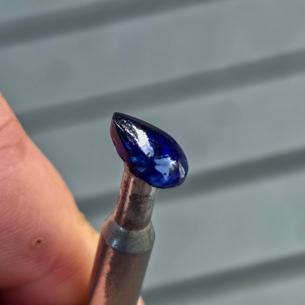 Close-up of a blue Madagascar sapphire dopped onto a metal dop stick, showing the stone’s glossy polished surface and teardrop-like profile while held between fingers. This image demonstrates a real gemstone mounted for lapidary work, providing context for how the Magnetic Dop Stick Tray supports stable staging, epoxy dopping, and repeatable handling during faceting and cabochon cutting workflows.