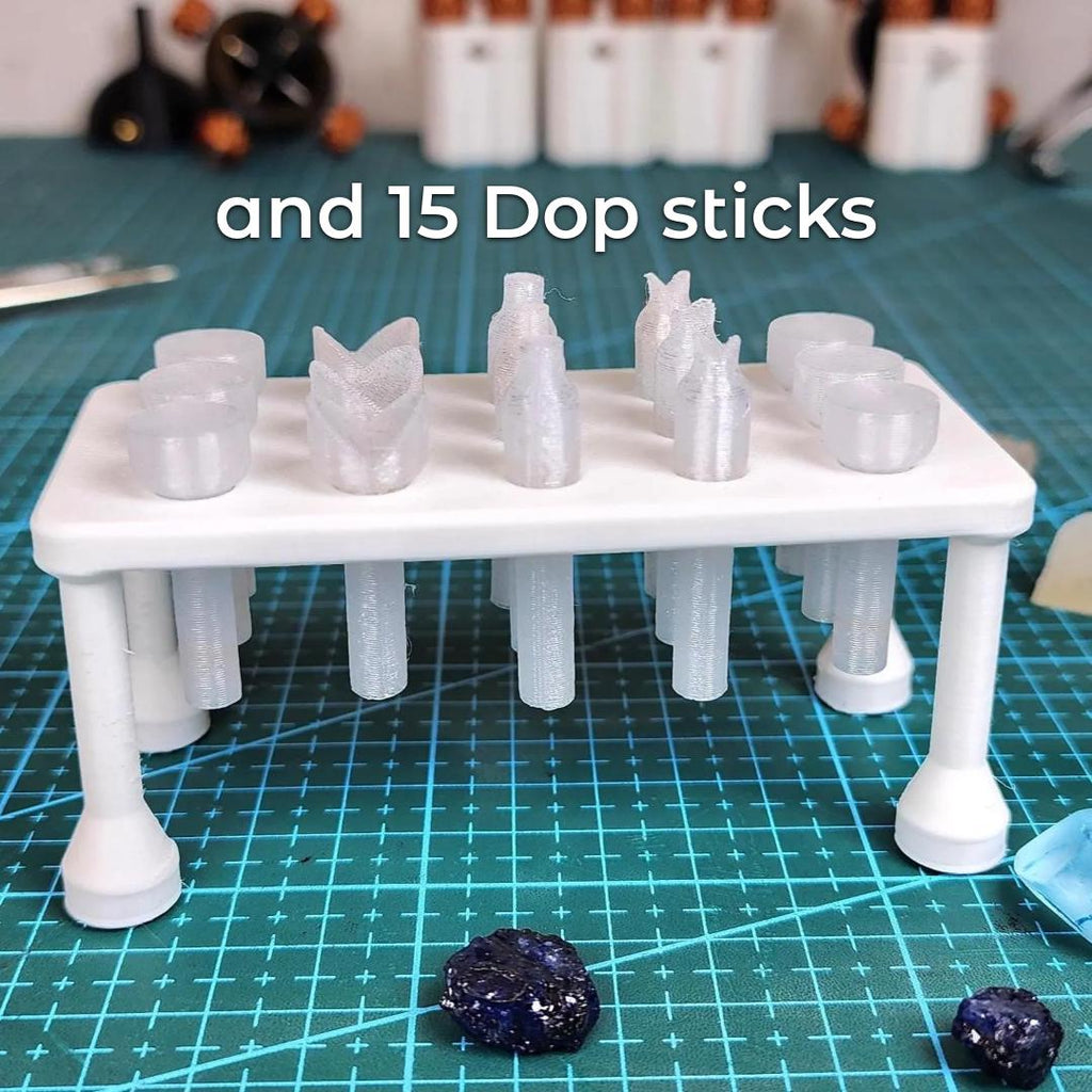 "A white Dop Stick Tray holding fifteen translucent 3D-printed dop sticks arranged neatly on a blue cutting mat. Various stone shapes and dop head styles are visible, showing customization options for different gem sizes. Raw gemstones rest nearby, illustrating real-world use for lapidary work and epoxy setting."