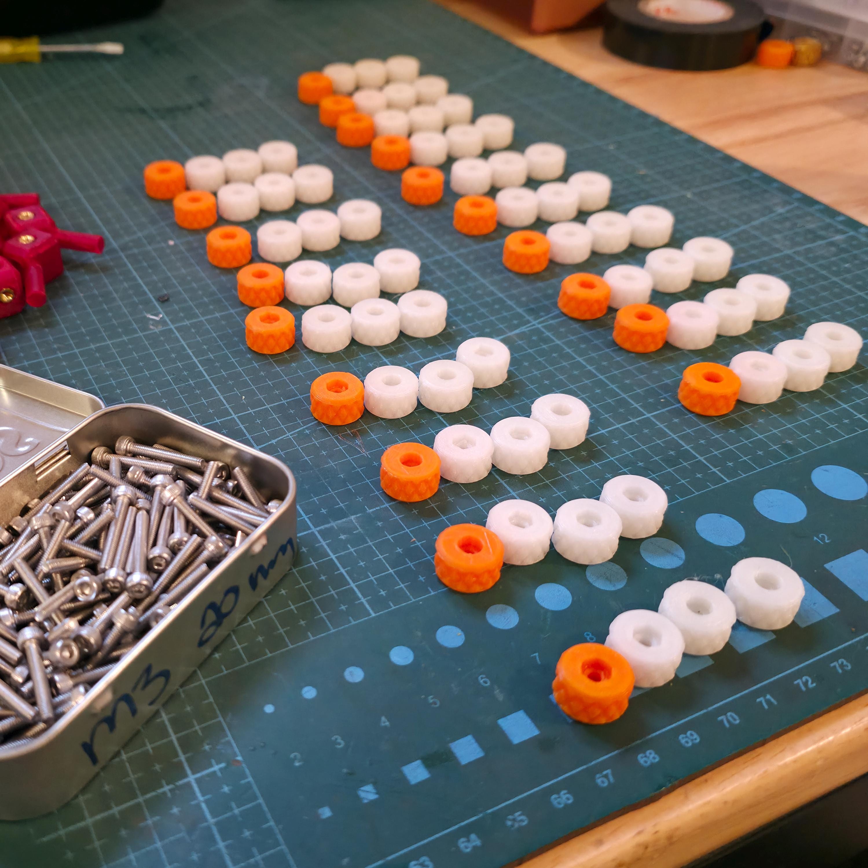 Organized rows of white and orange 3D-printed thumb nuts on a gridded cutting mat, beside a tin of M3 bolts. Demonstrates small-batch production, hardware kitting, and rapid manufacturing runs for custom components and prototype assemblies.