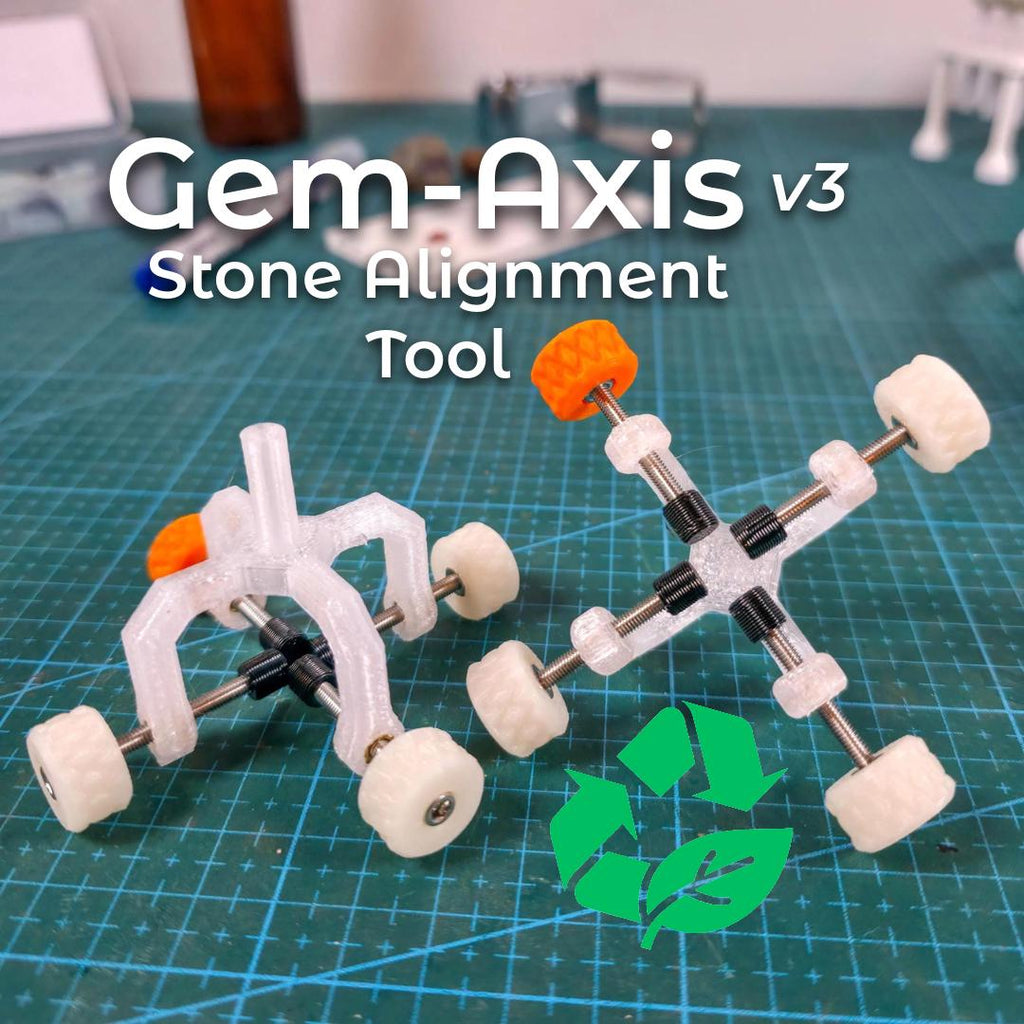 Two Gem Axis V3 stone alignment jigs on a green cutting mat in a bench setting. Clear recycled-polycarbonate bodies with stainless rods, white knurled wheels, and bright orange high-vis knobs. A green recycle-leaf icon appears at lower right. On-image text: “Gem-Axis v3 • Stone Alignment Tool.” Scene conveys precision centering at the dop to reduce epoxy drift and carat loss.
