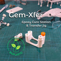 Aetherium Innovations Gem Xfer Magnetic Transfer Jig shown assembled with the Gem Axis Alignment Tool on a green cutting mat. The image highlights the fine-tune adjustment screws, alignment rods, and vertical epoxy-transfer configuration used for gemstone faceting.