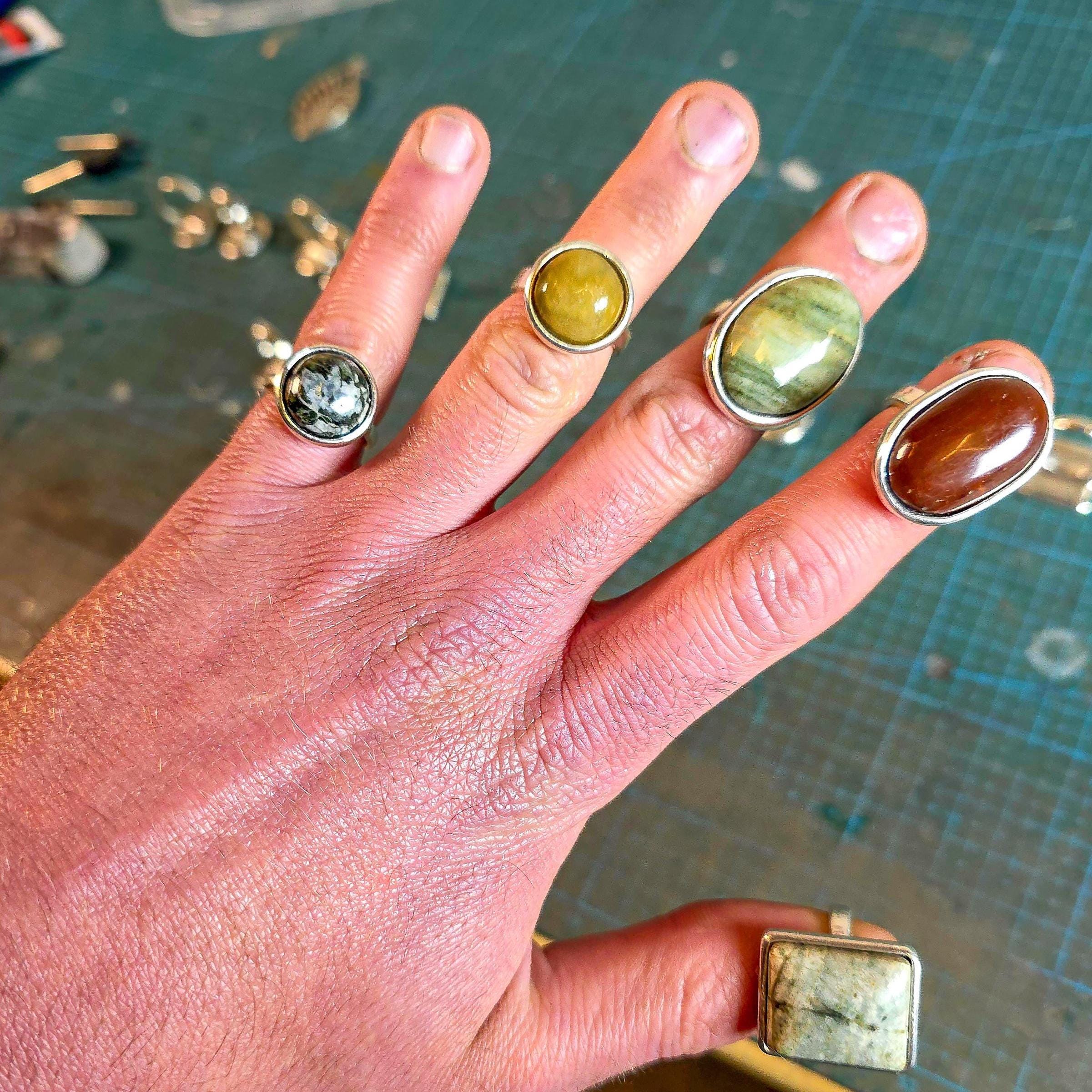 Close-up of a hand wearing multiple finished cabochon rings with polished stones. Natural stones in green, amber, yellow, and black tones. Highlights final fit and aesthetic results from the printed ring blanks after casting and polishing.