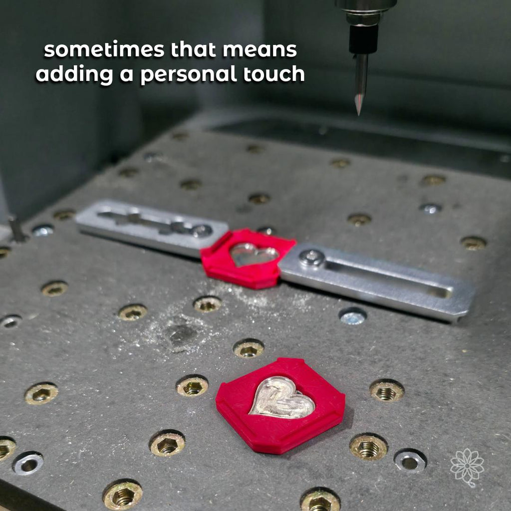 CNC milling machine carving details into a silver heart pendant held within a red custom fixture. Metal chips surround the workspace, emphasizing precision machining and hybrid-making techniques. Shows how fine engraving and personal elements are added to custom jewelry pieces.