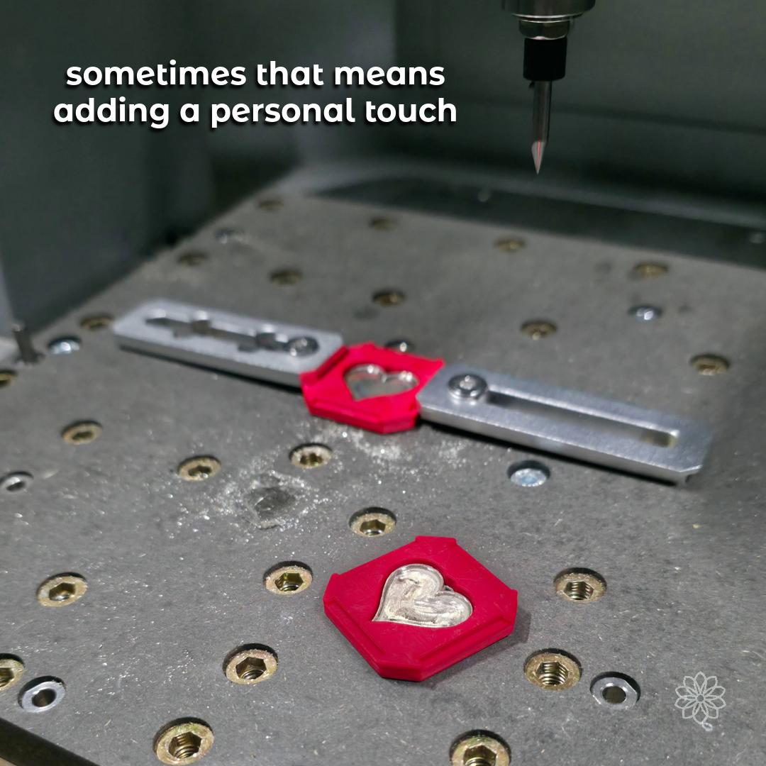 CNC milling machine carving details into a silver heart pendant held within a red custom fixture. Metal chips surround the workspace, emphasizing precision machining and hybrid-making techniques. Shows how fine engraving and personal elements are added to custom jewelry pieces.