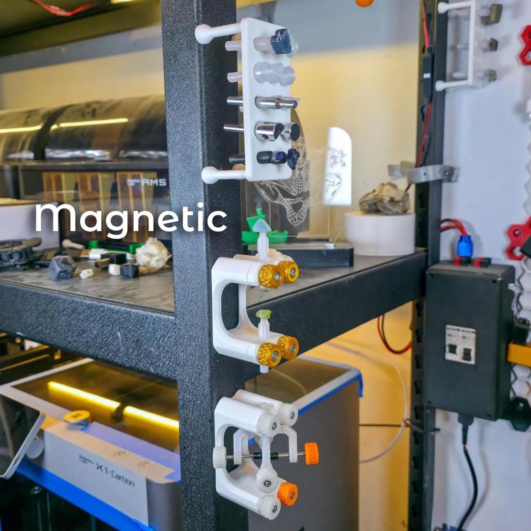"Gem Xfer and Dop Stick Tray tools magnetically attached to a black metal workbench support in a workshop. Several dop sticks with rough gemstones are mounted vertically for curing or storage. The image demonstrates the kit’s magnetic mounting capability and space-efficient organization for studio use."