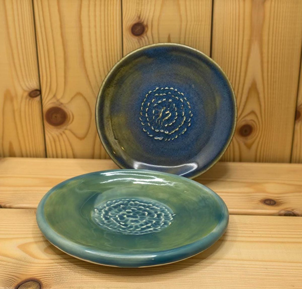 A blue ceramic plate with a raised spiral texture stands upright against wooden boards, while a green-toned plate rests flat below it. Both plates have glossy handmade finishes, and the warm wood background enhances their color depth and artisan detail.