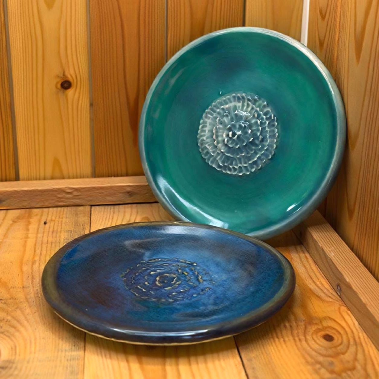 Two handcrafted ceramic plates displayed inside a wooden nook. The top plate has a green and yellow glaze with a textured center, while the lower plate shows blue tones. Light and shadow across the wood emphasize the plates’ glossy surfaces and soft color transitions.