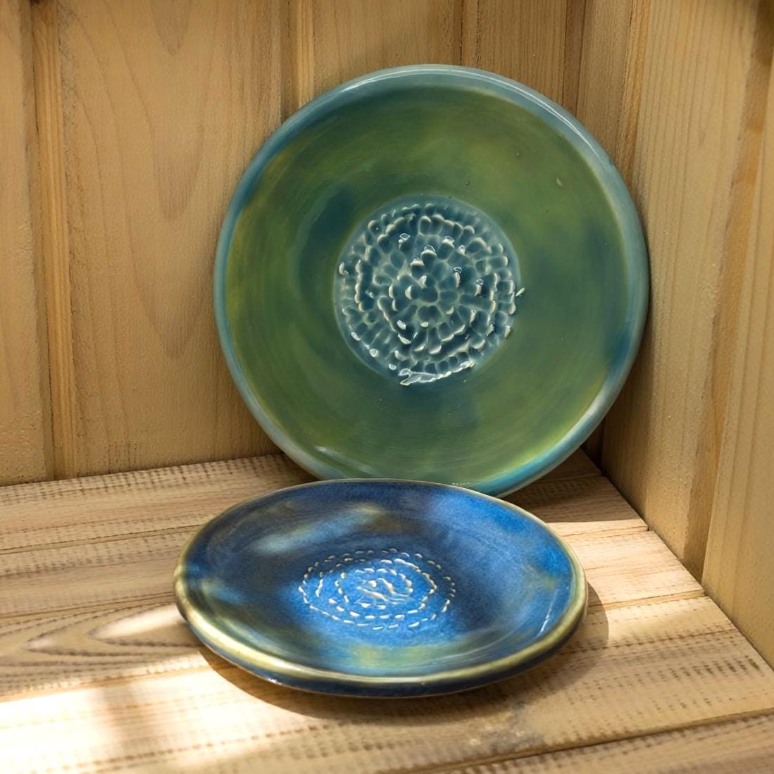 Two ceramic plates in teal and deep blue glazes are positioned against wooden walls. The raised spiral centers add texture, while the glossy surfaces reflect ambient light. The warm wood tones frame the handmade pieces and highlight their color depth.